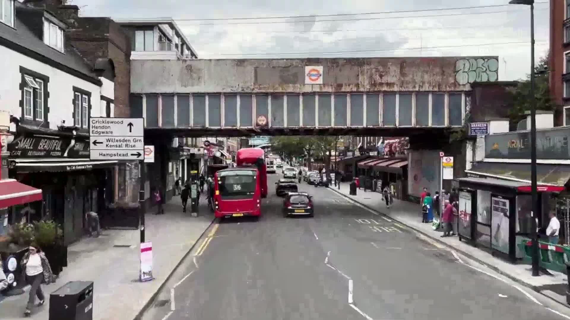 A street scene shows two red double-decker buses and several cars driving under a railway bridge on an overcast day, with pedestrians walking along the sidewalks and shops lining the street.