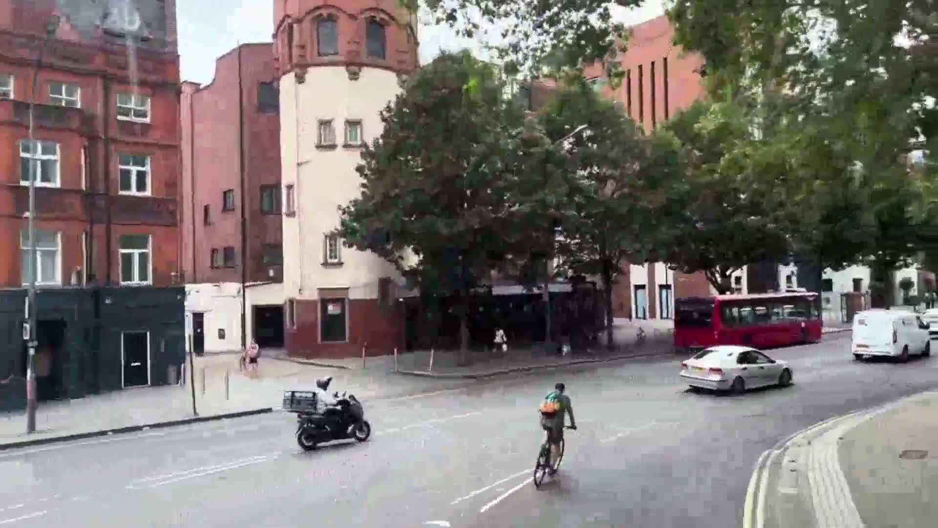 A city street scene on an overcast day shows a red bus, several cars, a motor scooter, and a bicycle, all moving near brick buildings and lush green trees.