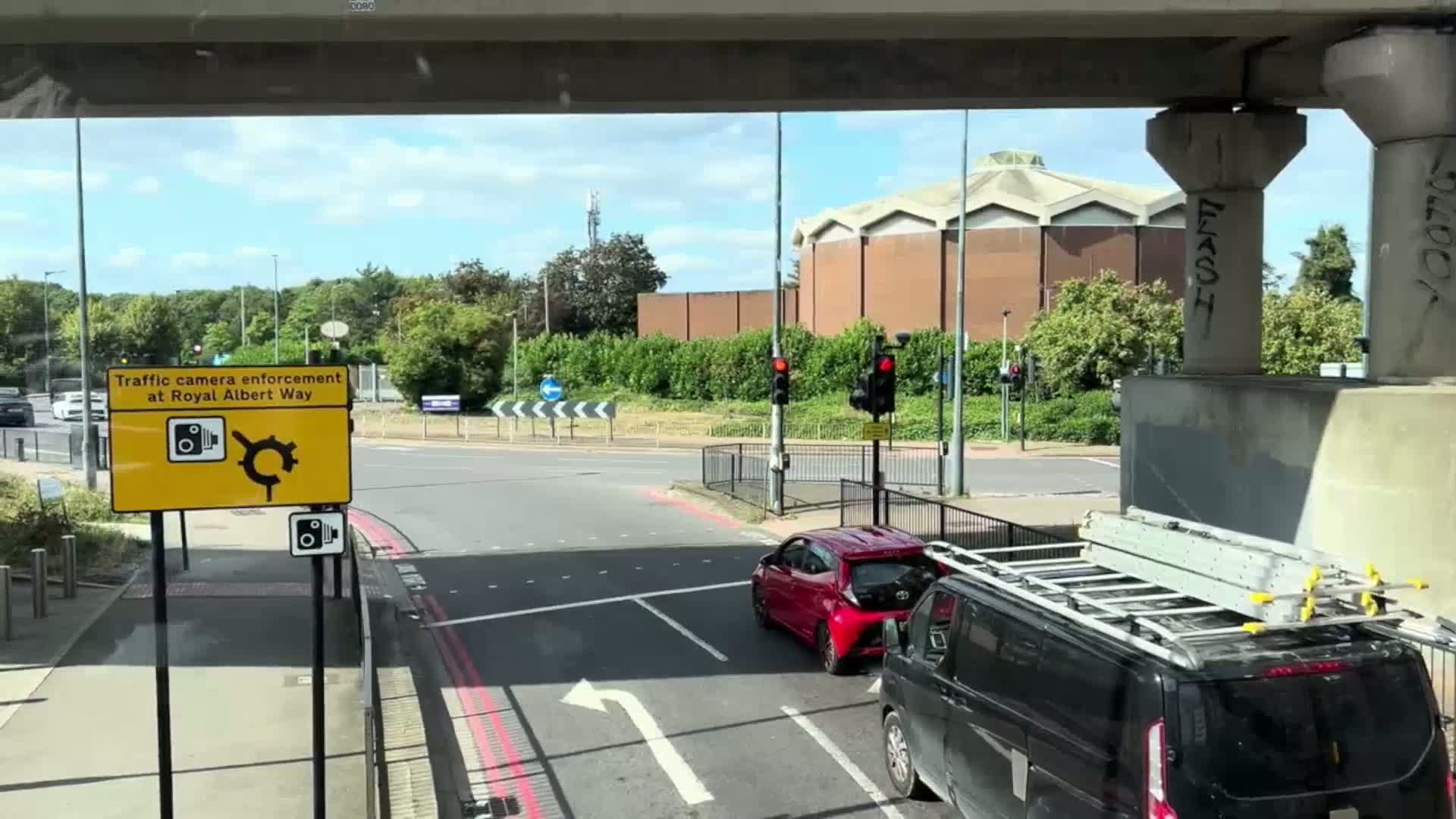 A view from under a bridge on a sunny day shows a roundabout with a red car and a black van with ladders on top approaching a traffic light.