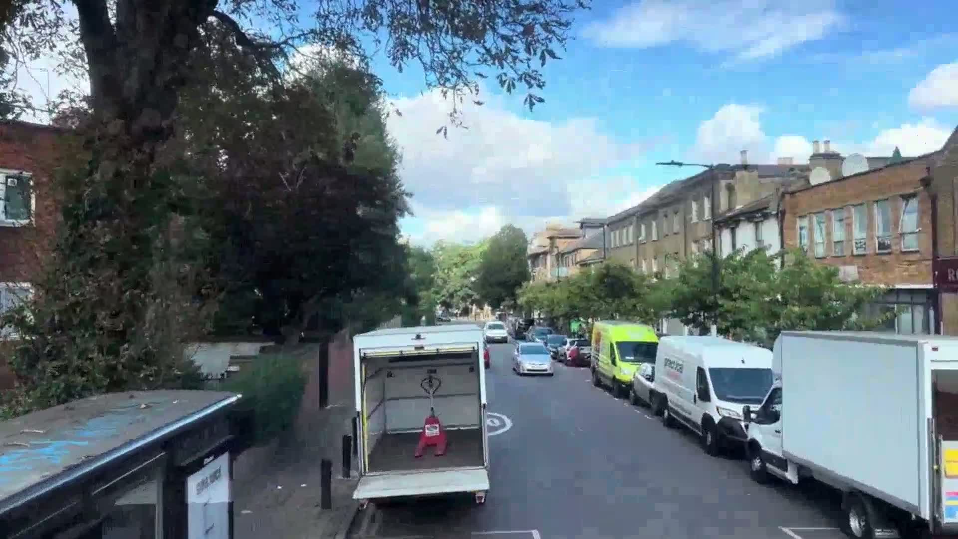 A partly sunny day shows a residential street with parked cars and vans, a white van with its rear door open revealing a pallet jack, and a bus stop with graffiti.