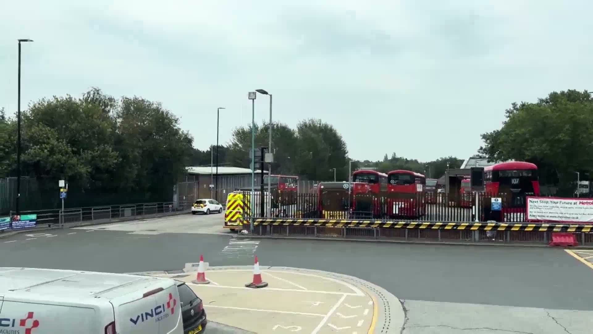 Several red double-decker buses are parked behind a fence in a bus depot on an overcast day.