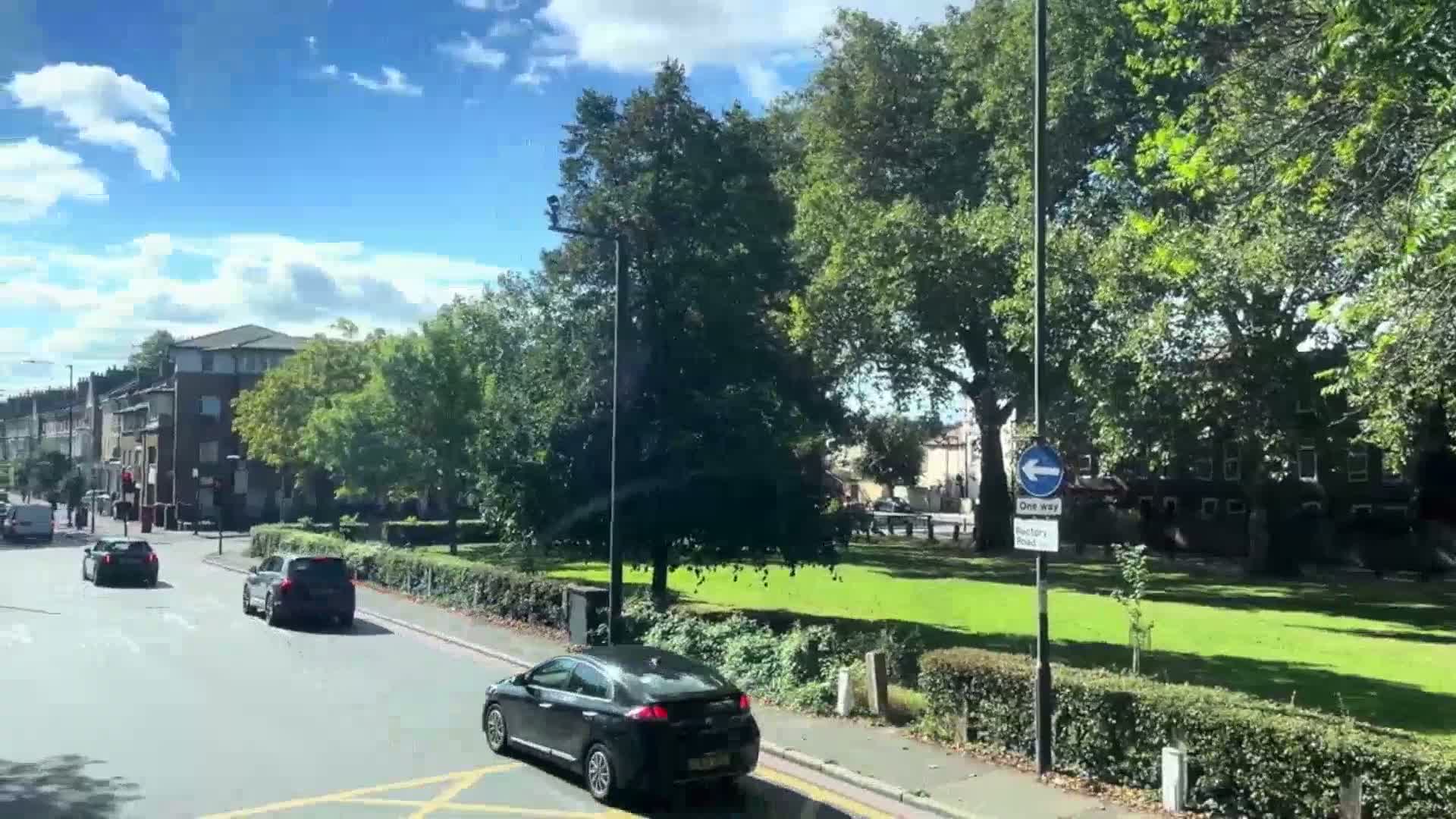 A sunny day reveals a residential street scene with several cars driving on a road that passes a grassy park bordered by lush green trees and hedges.