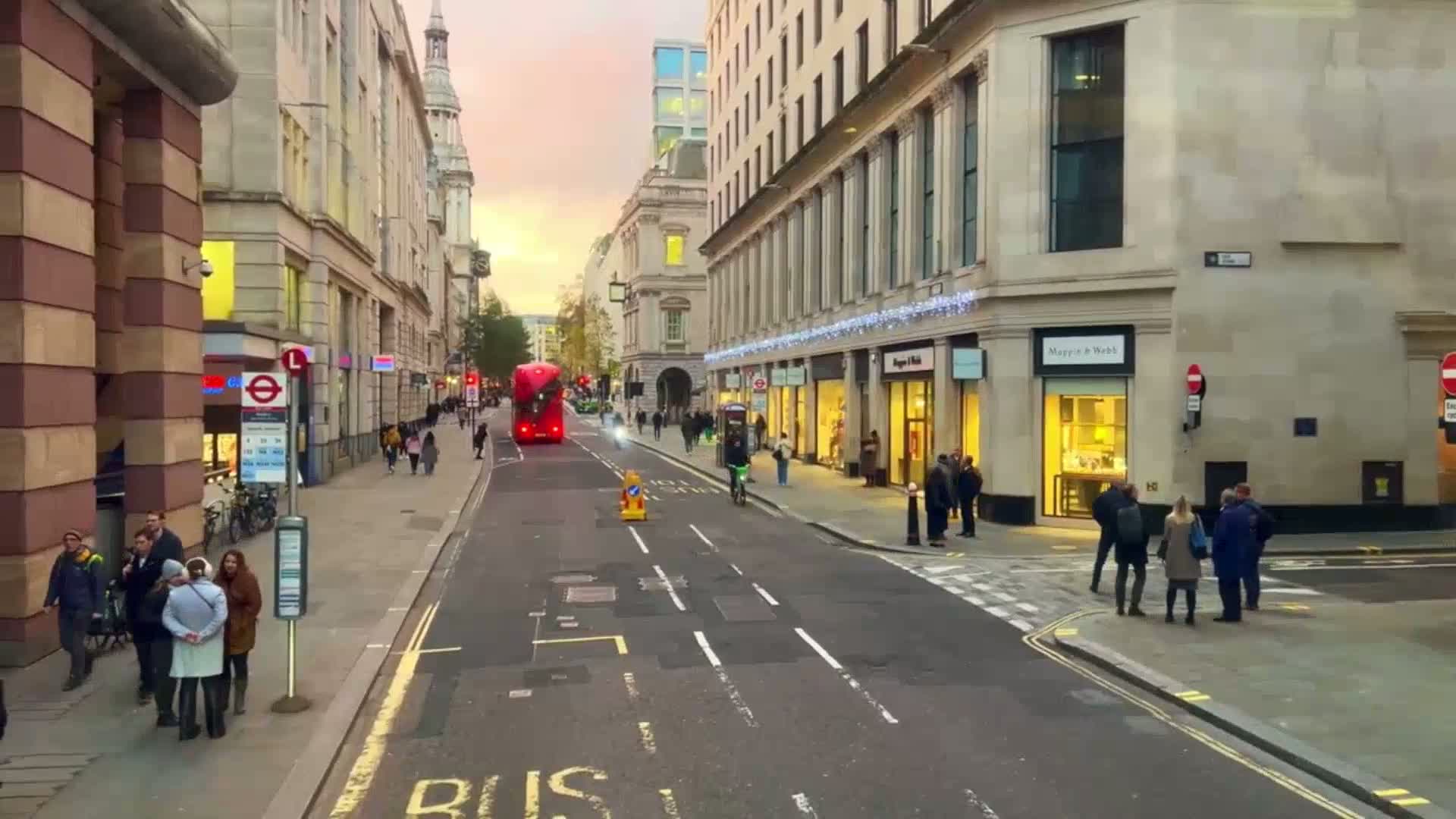 A city street at dusk, with pedestrians walking past shops and a red double-decker bus driving down the road.