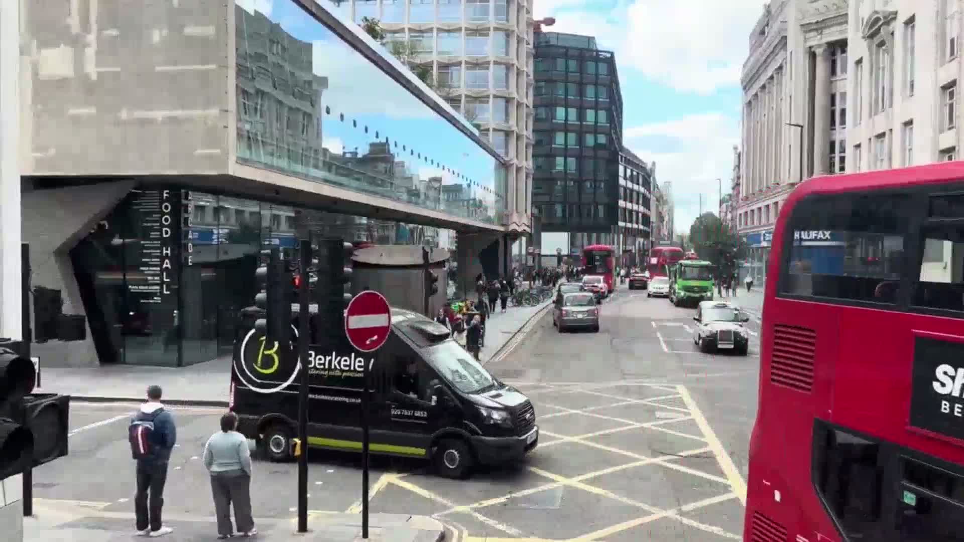 A city street scene on a sunny day shows several vehicles at a crossroads, pedestrians walking along a sidewalk, and modern buildings lining the street.