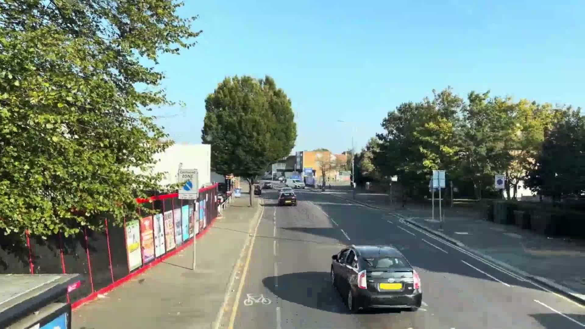 A sunny day shows a multi-lane road with several cars driving, lined with trees and buildings, and a construction fence with posters on the side.