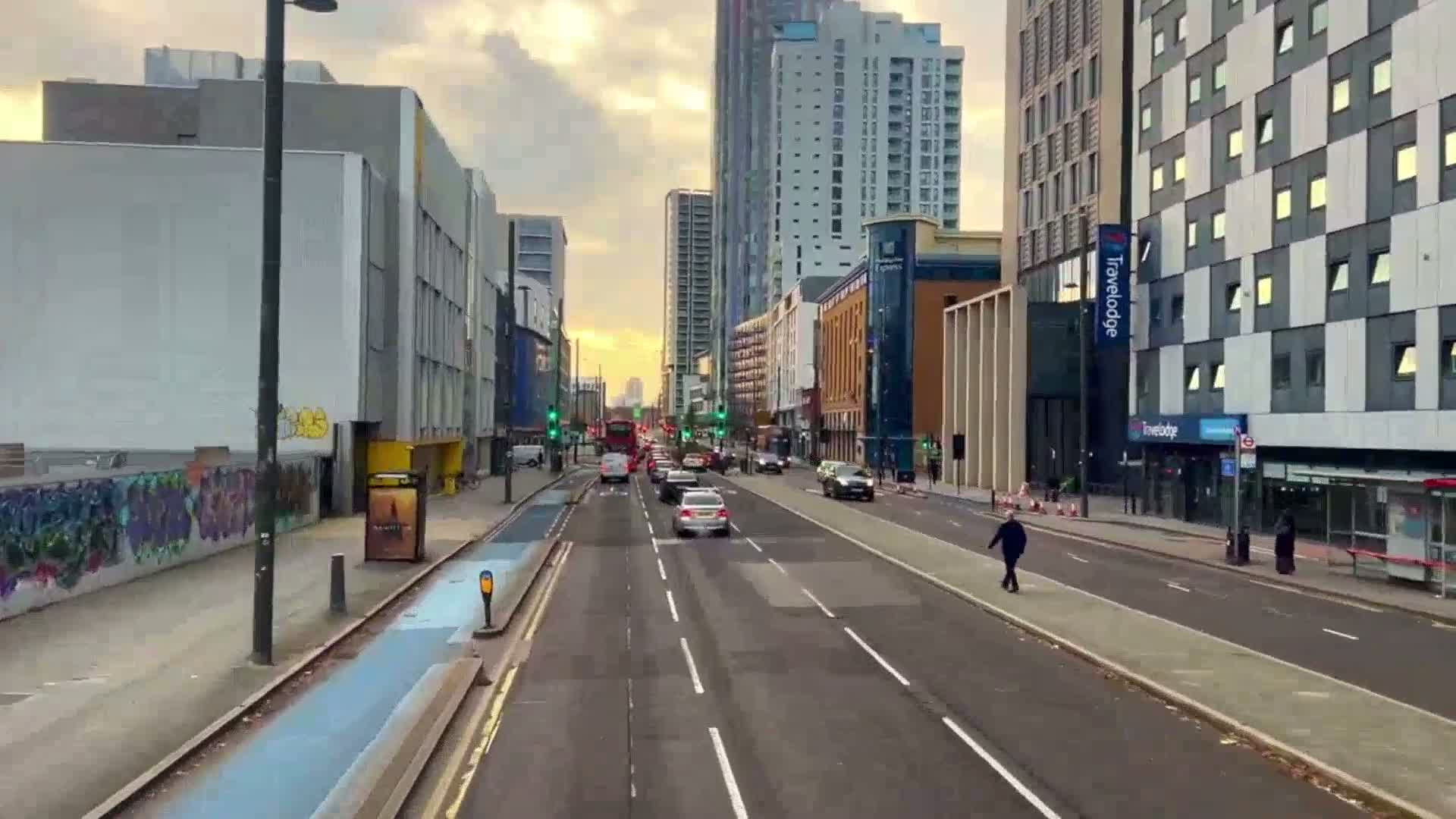 A city street with several lanes of traffic under a partly cloudy sunset sky, flanked by modern buildings and a graffiti-covered wall.