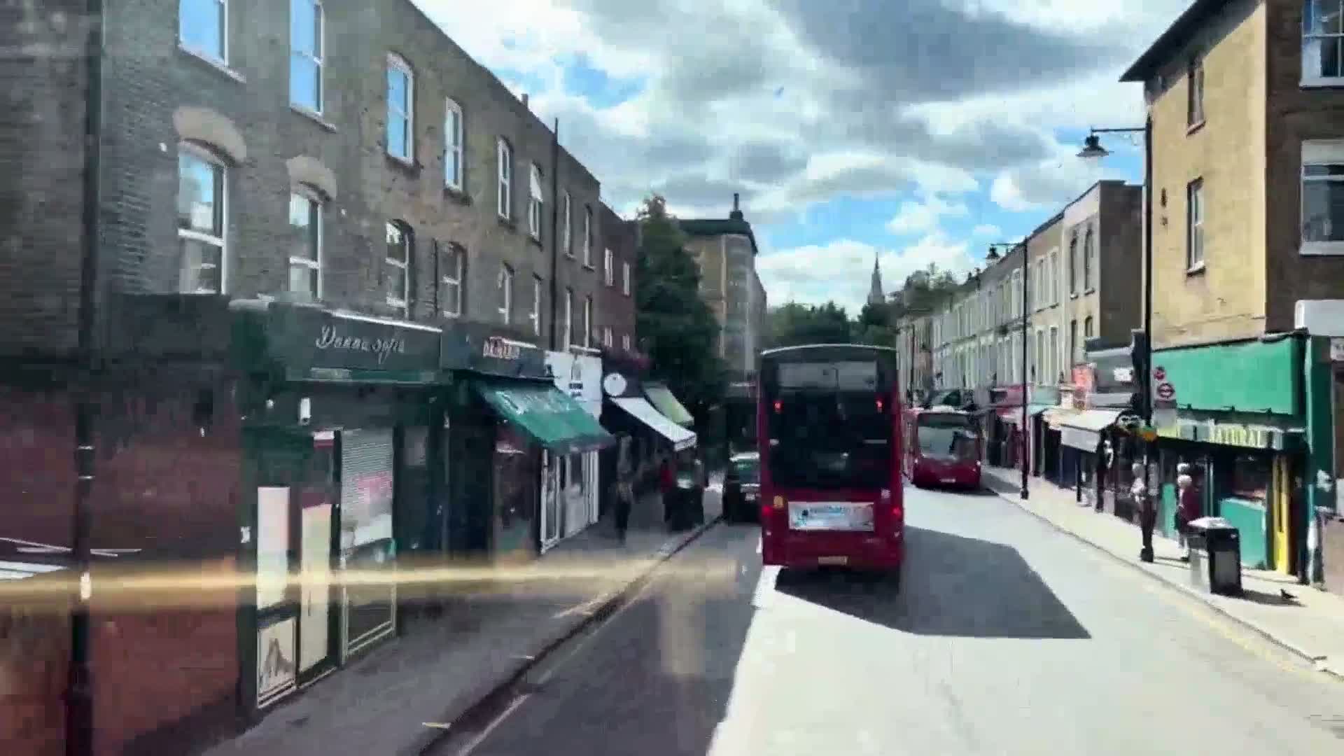 A partly sunny day shows a street lined with shops and buildings, with two double-decker buses driving down the road.