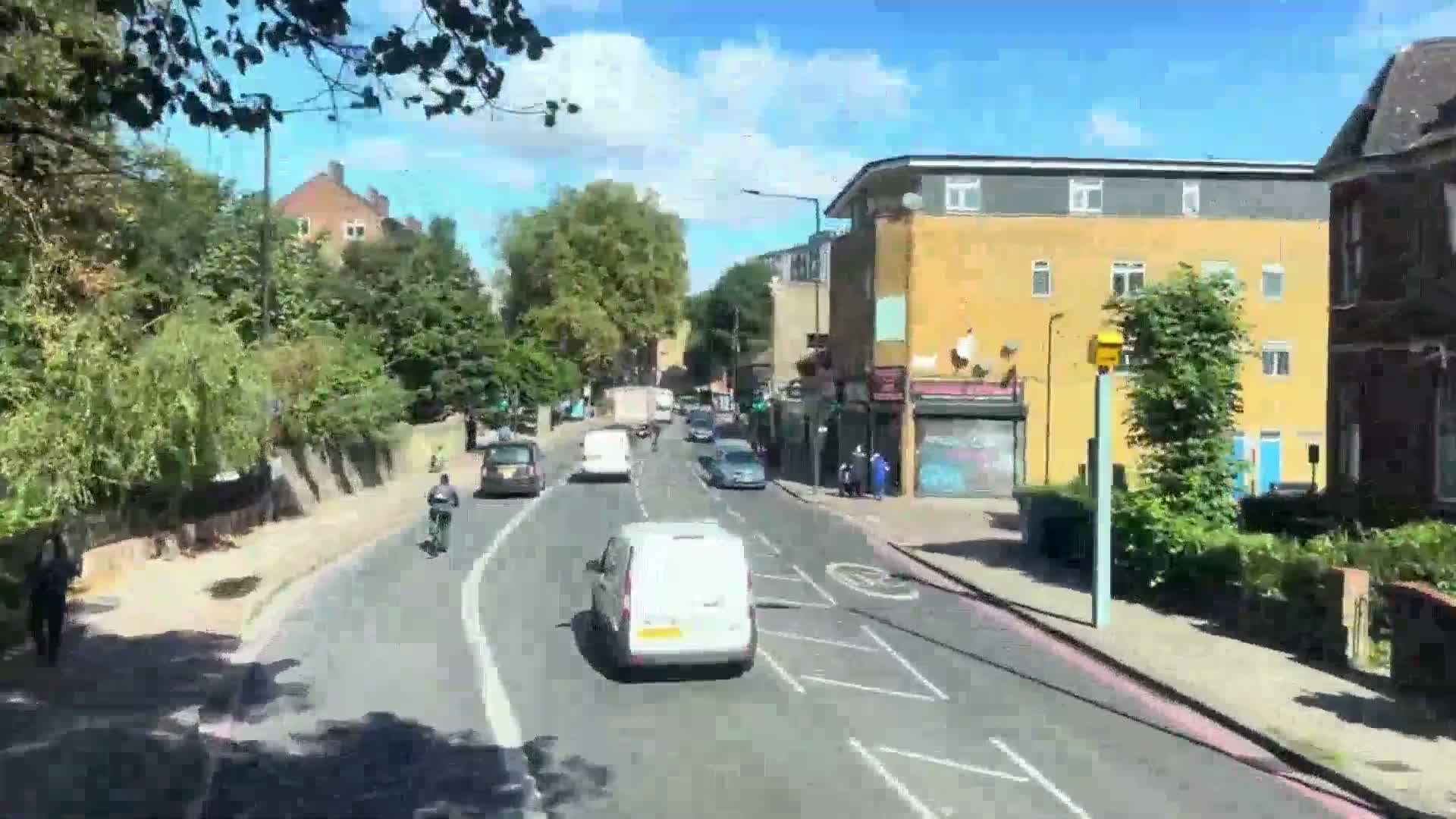 A sunny day shows a residential street with several cars and two cyclists traveling in the same direction, passing by shops and houses lined with green trees.