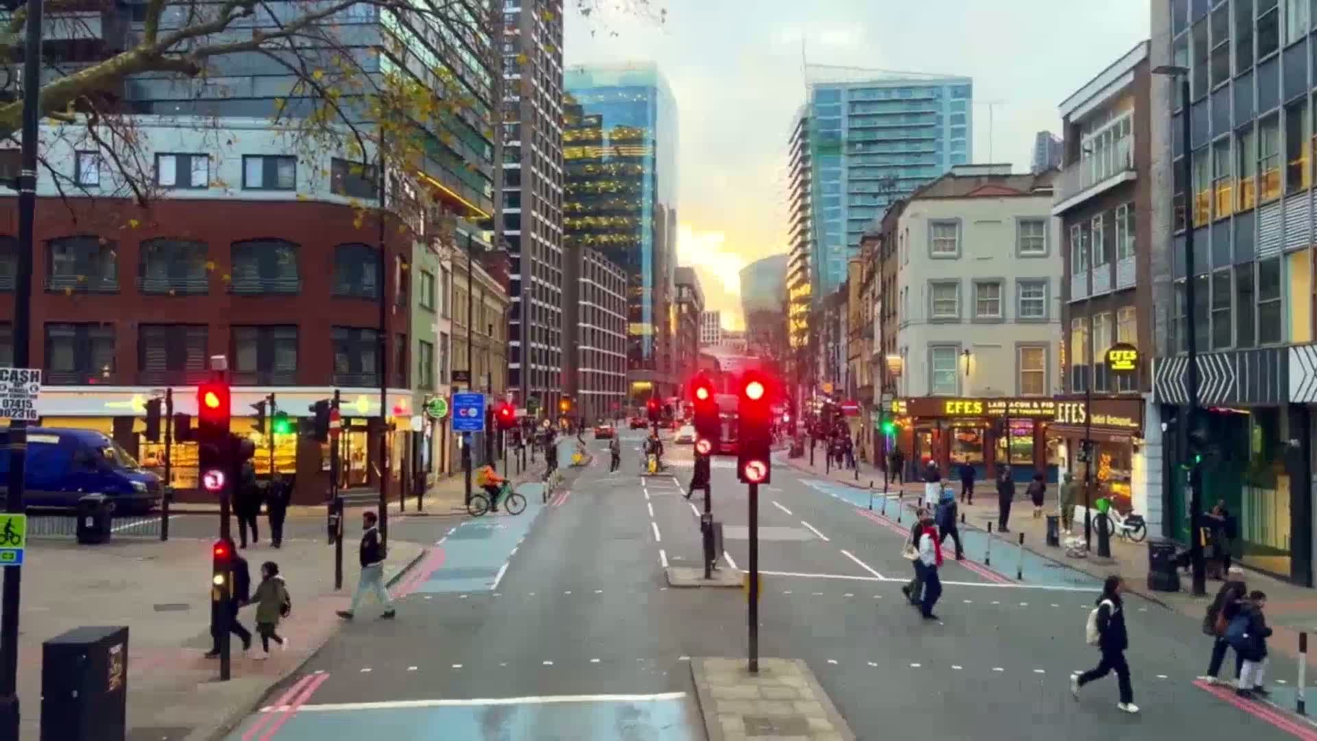 A city street scene at sunset shows pedestrians crossing a road lined with multi-story buildings, some with shops on the ground floor, under a mostly clear sky.