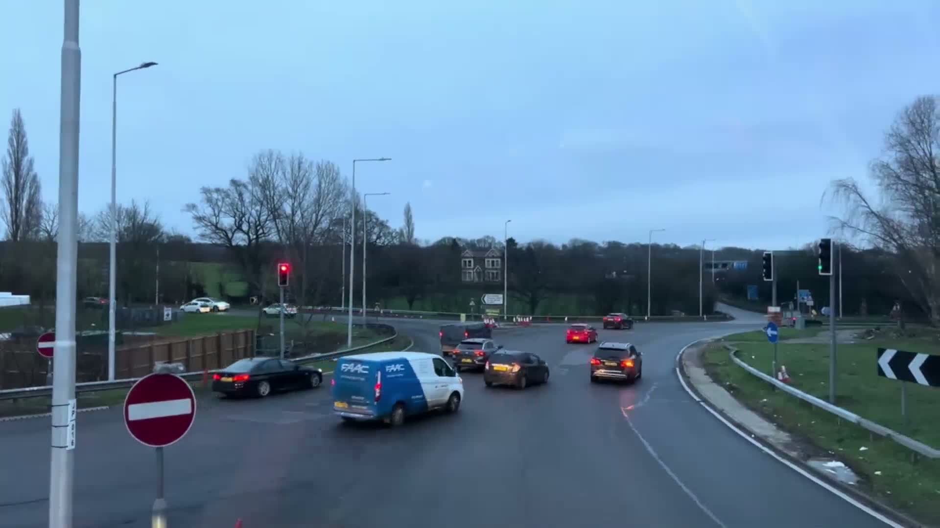 Several cars and a van are navigating a roundabout under an overcast sky.