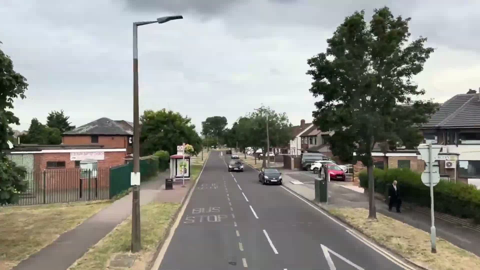 A residential street with overcast skies shows several cars driving and a man walking on the sidewalk, along with a bus stop and several houses.