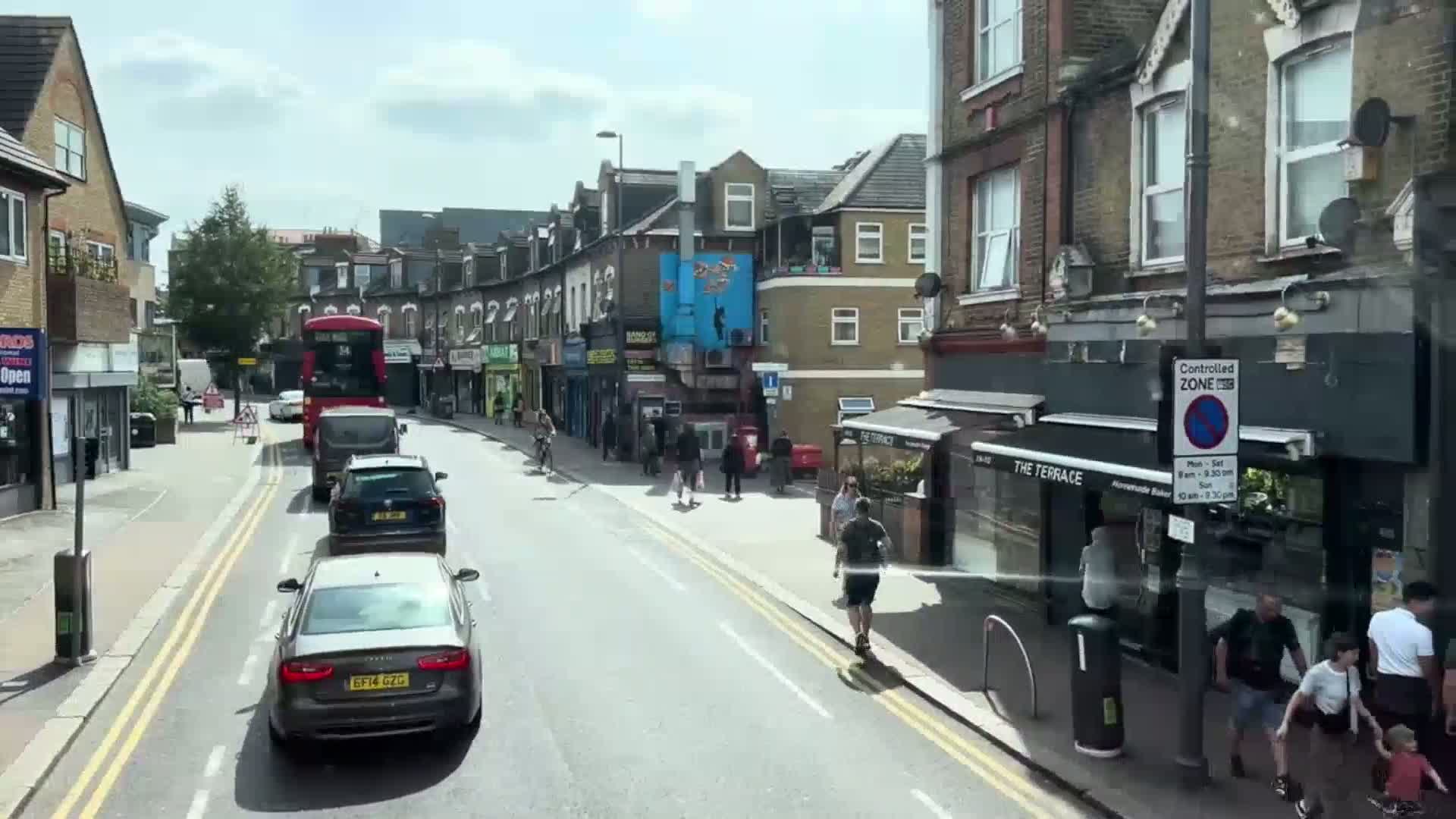 A sunny day on a residential street with several cars, pedestrians, and a double-decker bus in the distance.