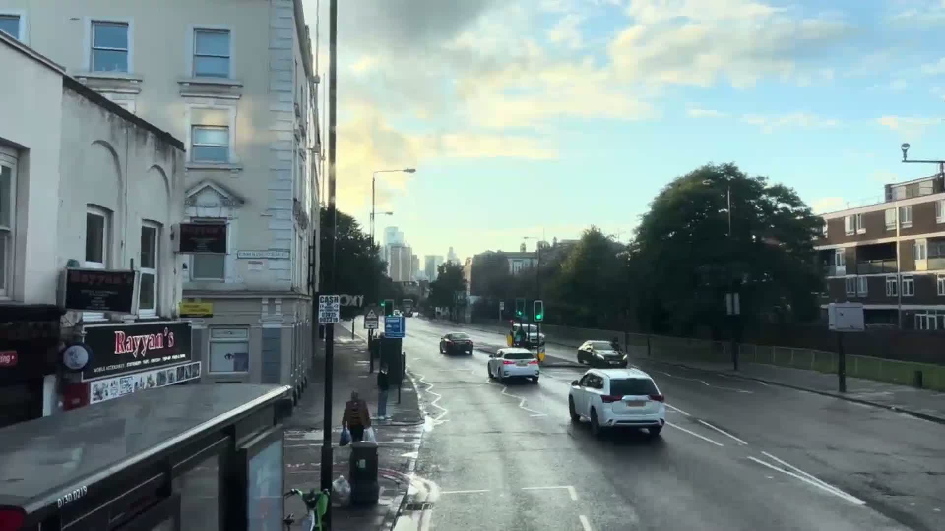 A wet street scene shows several cars driving on a road lined with buildings, under a partly cloudy sky.