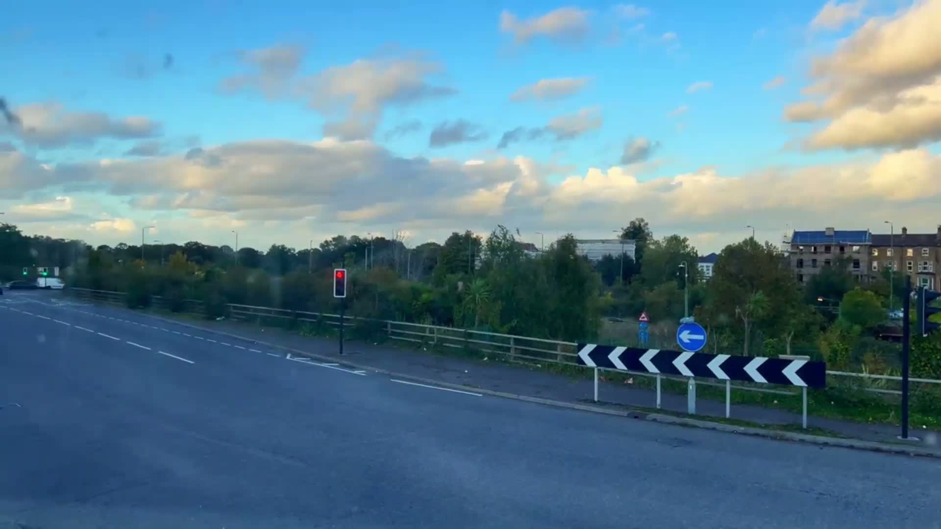 A mostly sunny day with scattered clouds shows a road intersection with a red traffic light and a blue directional sign, beyond which are trees and buildings.