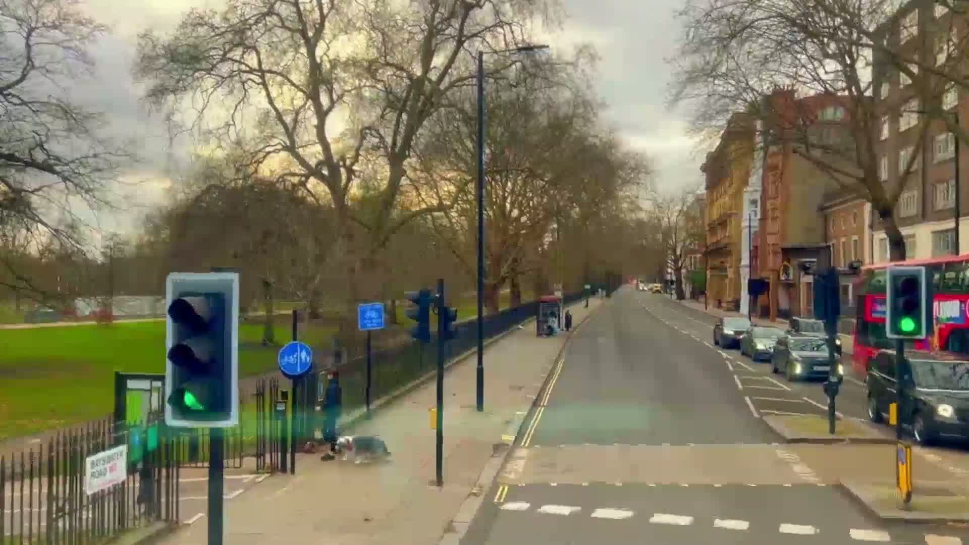 A city street scene on an overcast day shows a traffic light displaying green, several cars driving, pedestrians walking alongside a park, and a double-decker bus partially visible.