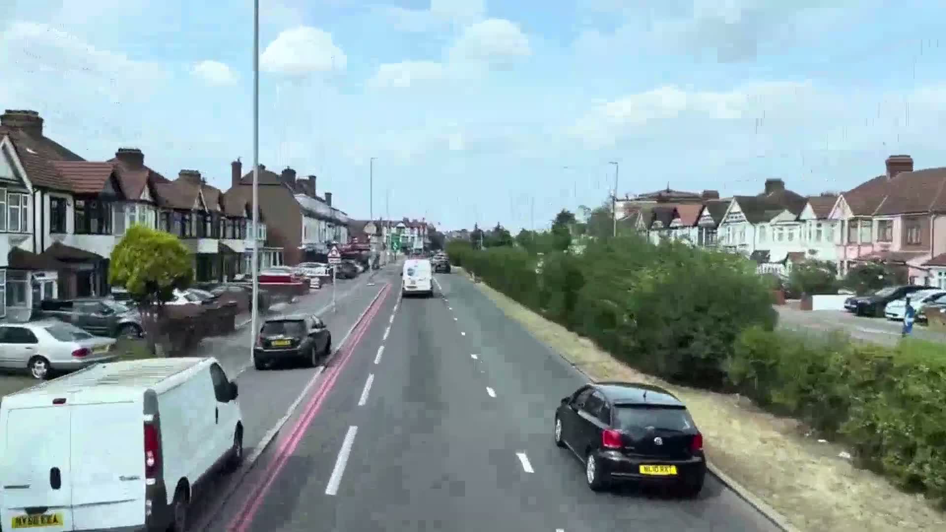 A sunny day reveals a residential street lined with houses, with cars driving on a road bordered by a hedge.