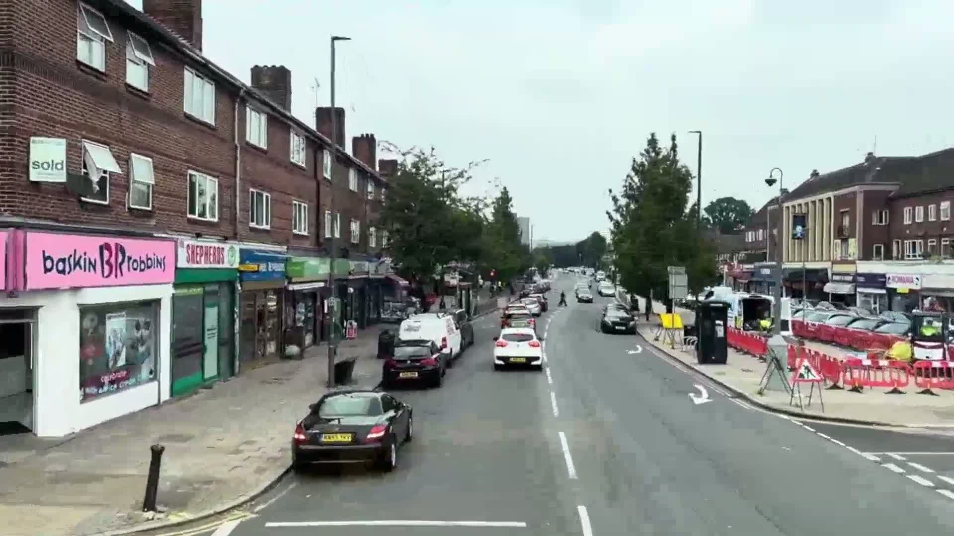 A street lined with shops and parked cars on an overcast day, with several cars driving down the road.