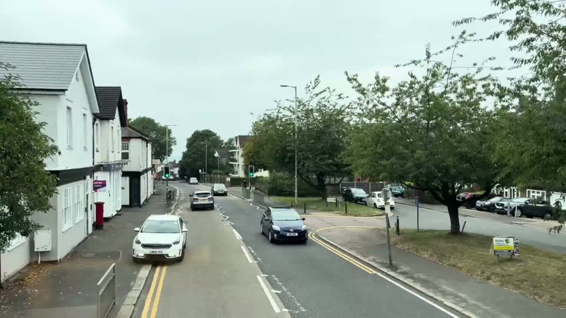 A street scene on an overcast day shows several cars driving past two-story buildings and leafy green trees.