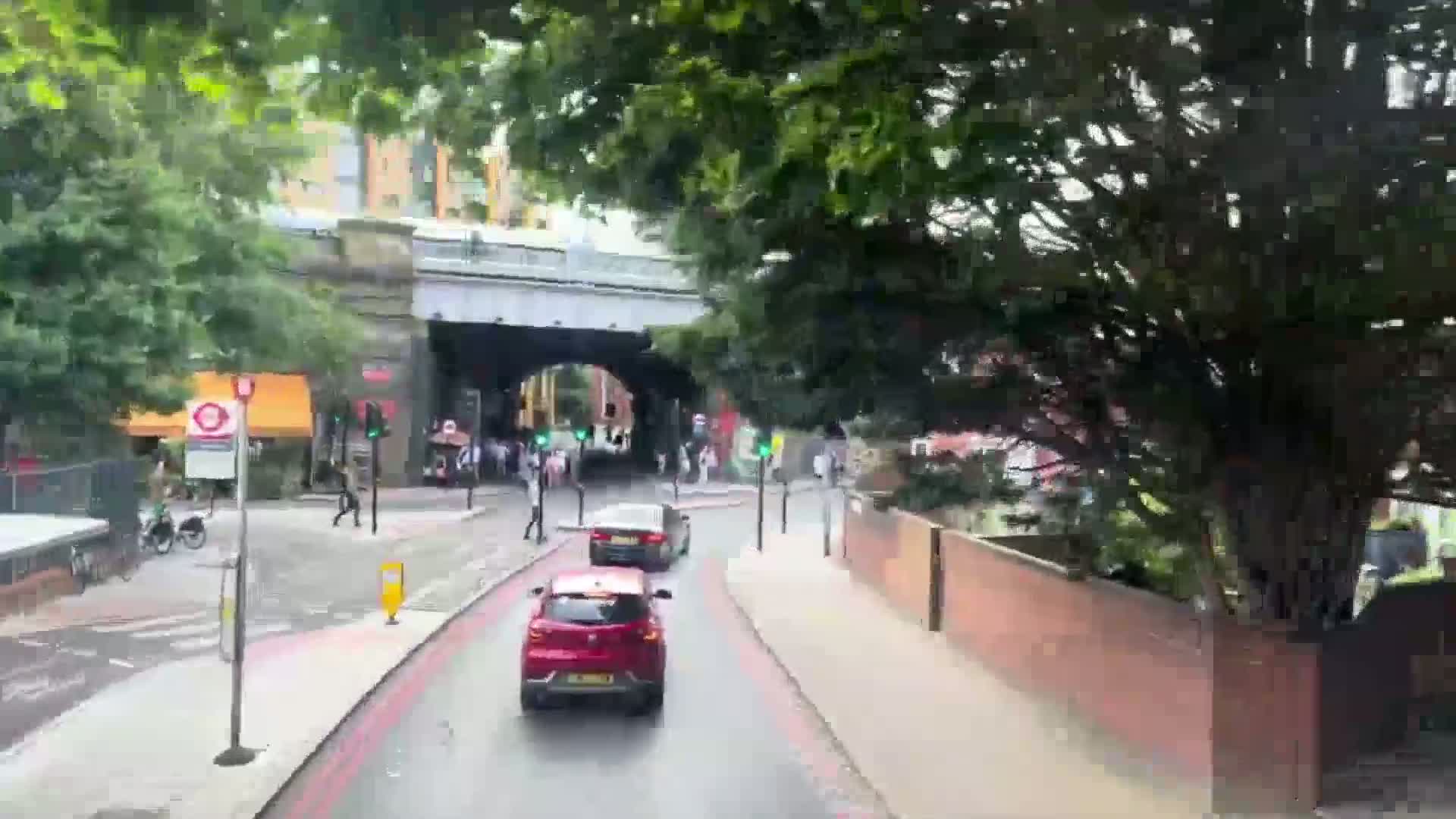 A street scene on an overcast day shows several cars driving past two-story buildings and leafy green trees.