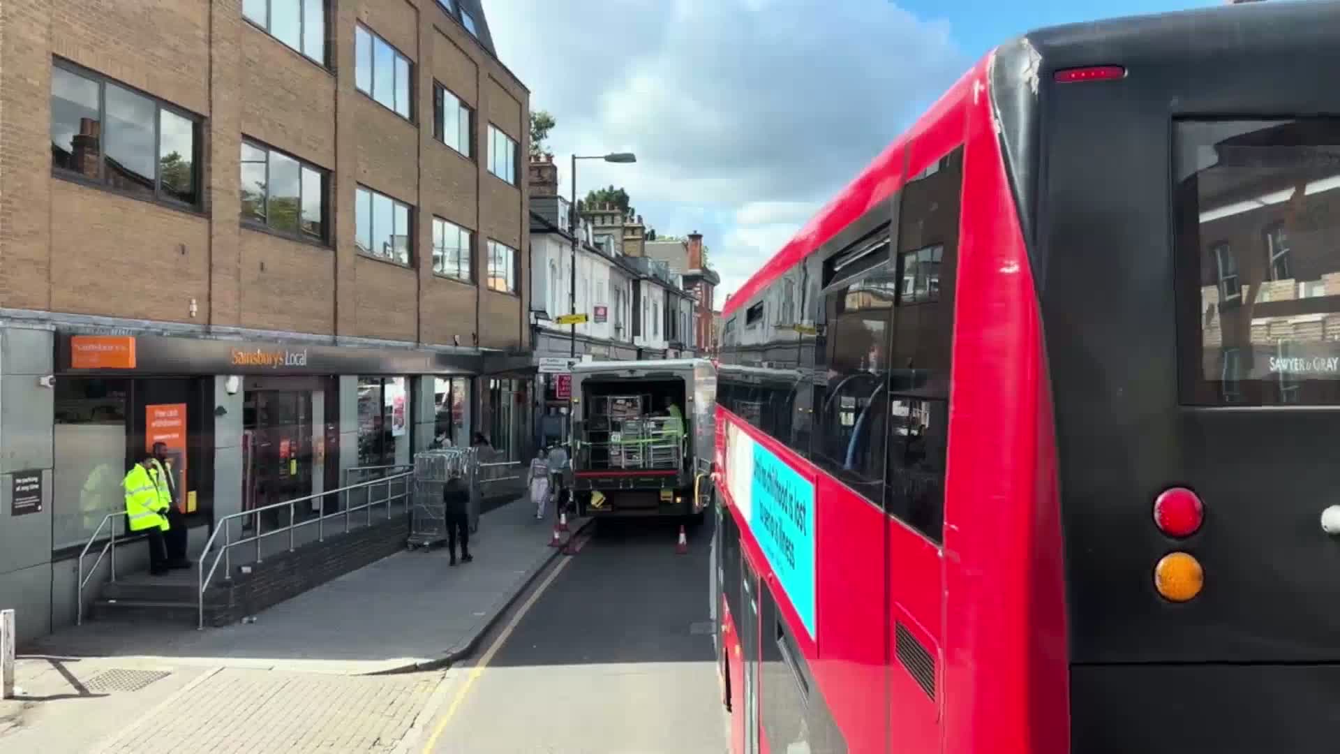 A red double-decker bus is stopped on a city street on a partly cloudy day, behind a delivery truck unloading goods in front of a Sainsbury's Local store.