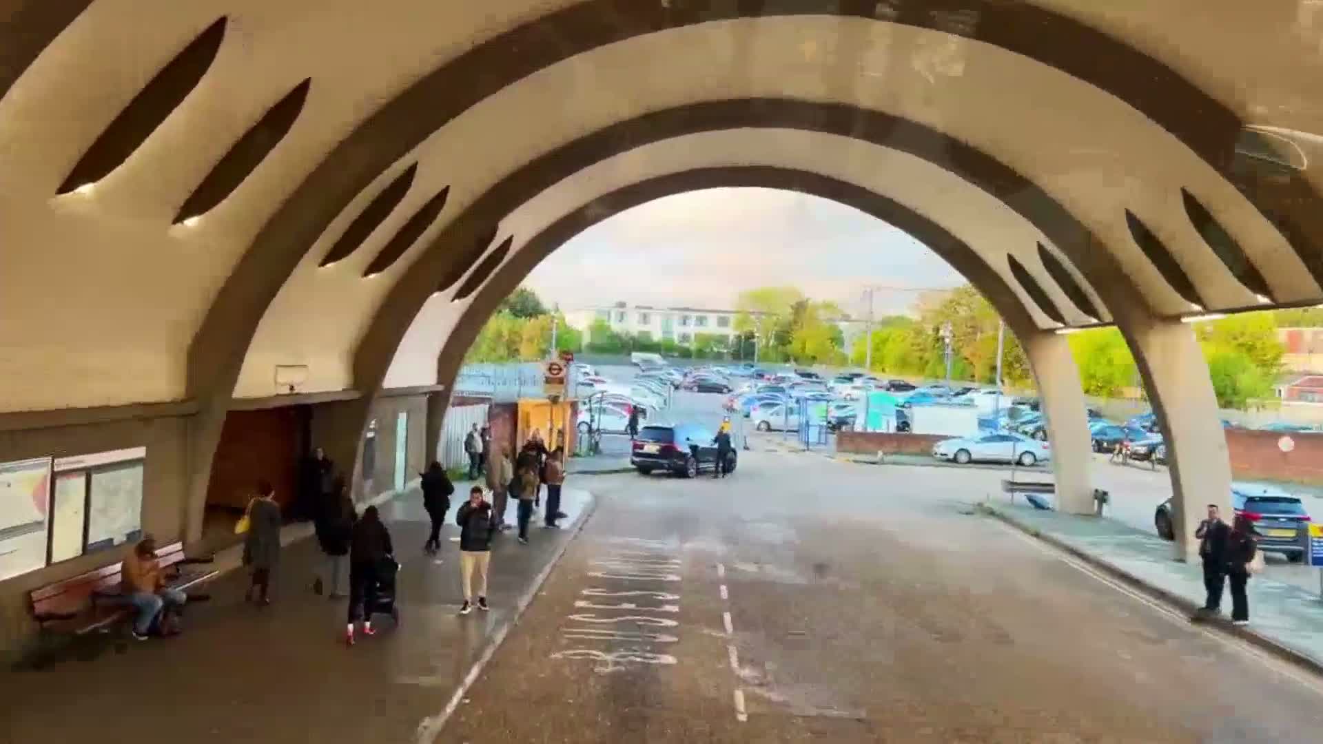 A view from inside a curved bus shelter shows several people waiting outside, a parking lot in the background under an overcast sky.