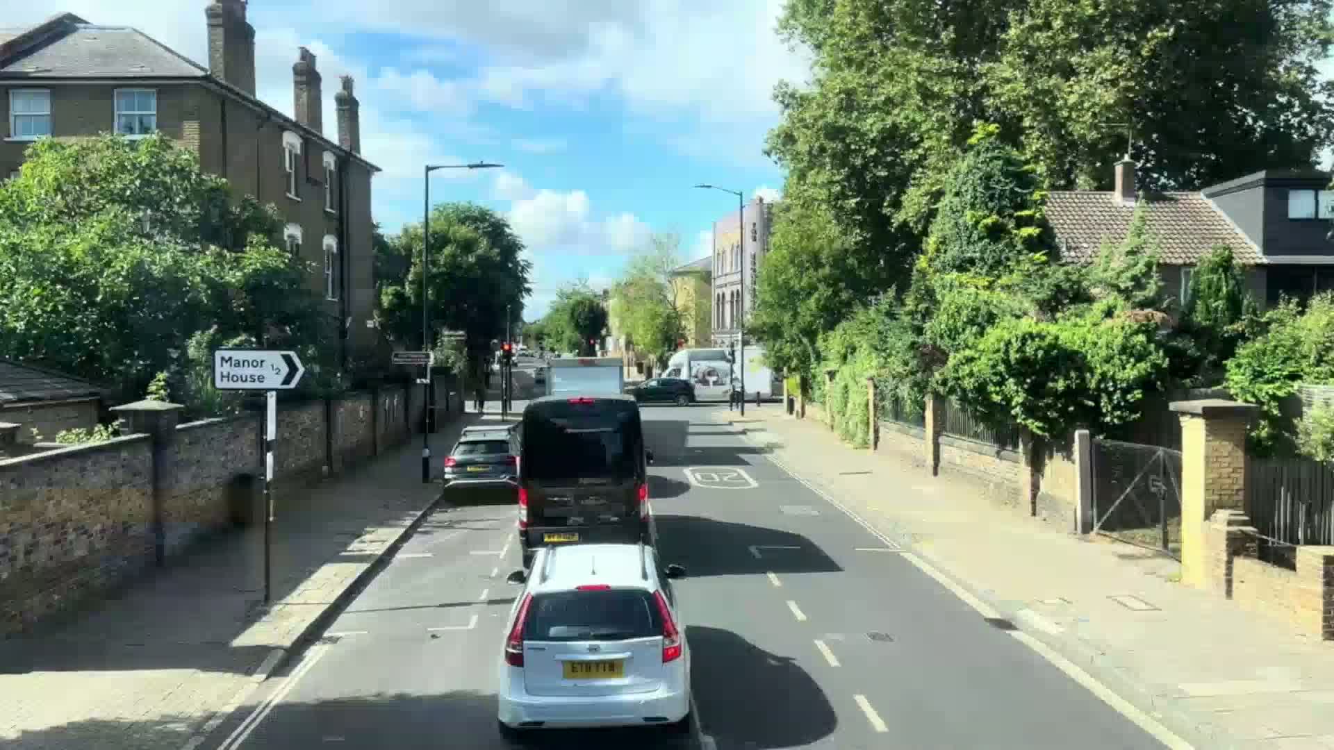A sunny day reveals a residential street with several cars driving in both directions, lined with brick houses and lush green trees.