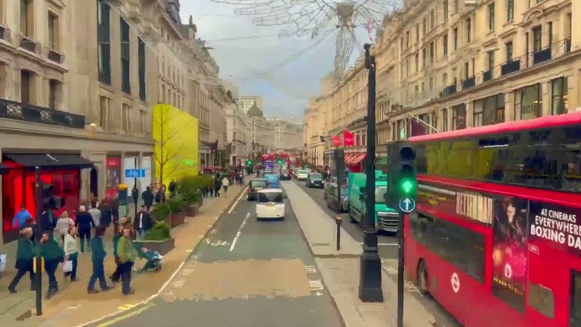 A city street scene on an overcast day shows pedestrians walking along a sidewalk lined with shops and buildings, while cars and a double-decker bus travel down the road.
