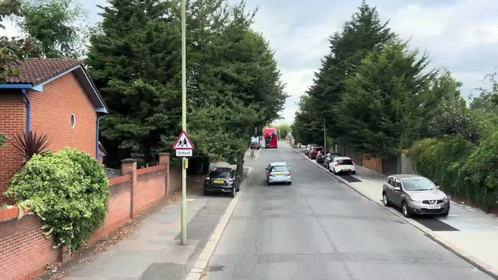 A residential street lined with cars, brick houses, and lush green trees under an overcast sky shows a red double-decker bus in the distance.
