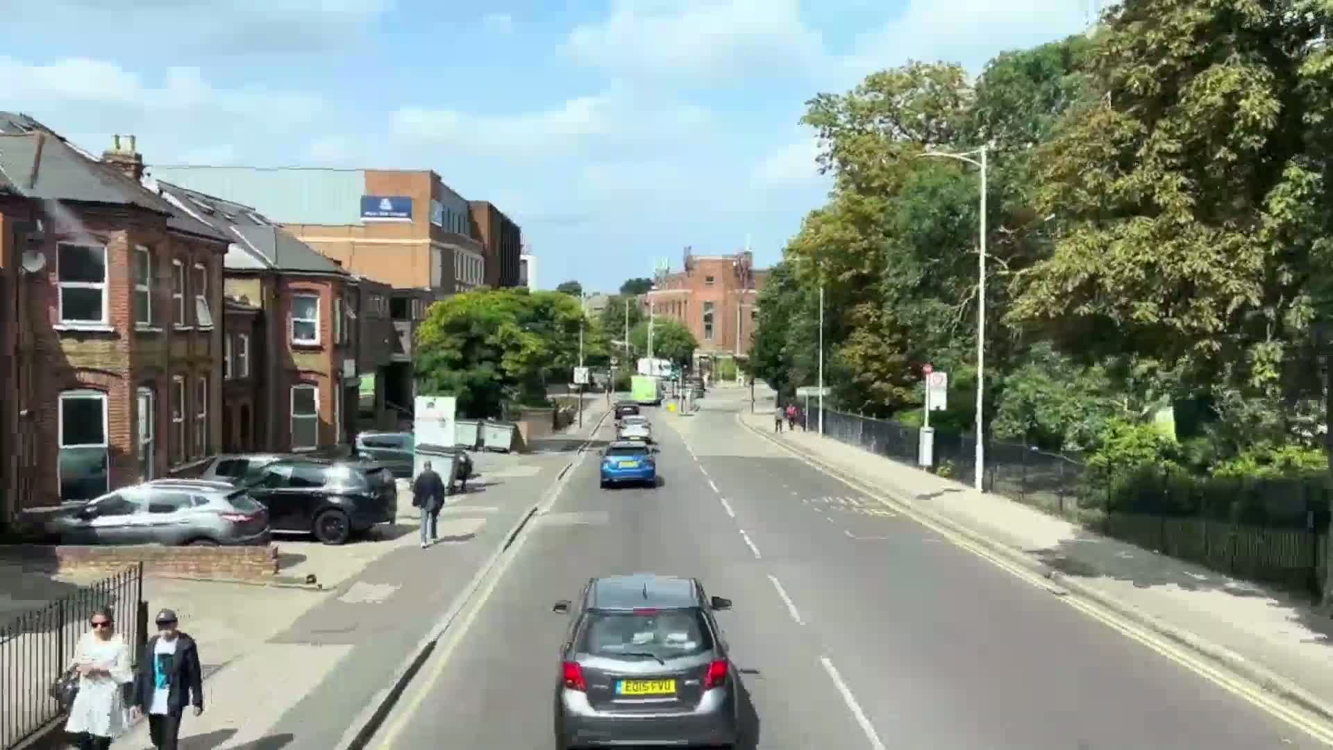 A sunny day reveals a tree-lined street with parked cars along the sides and several cars driving down the road, past brick buildings and lush greenery.