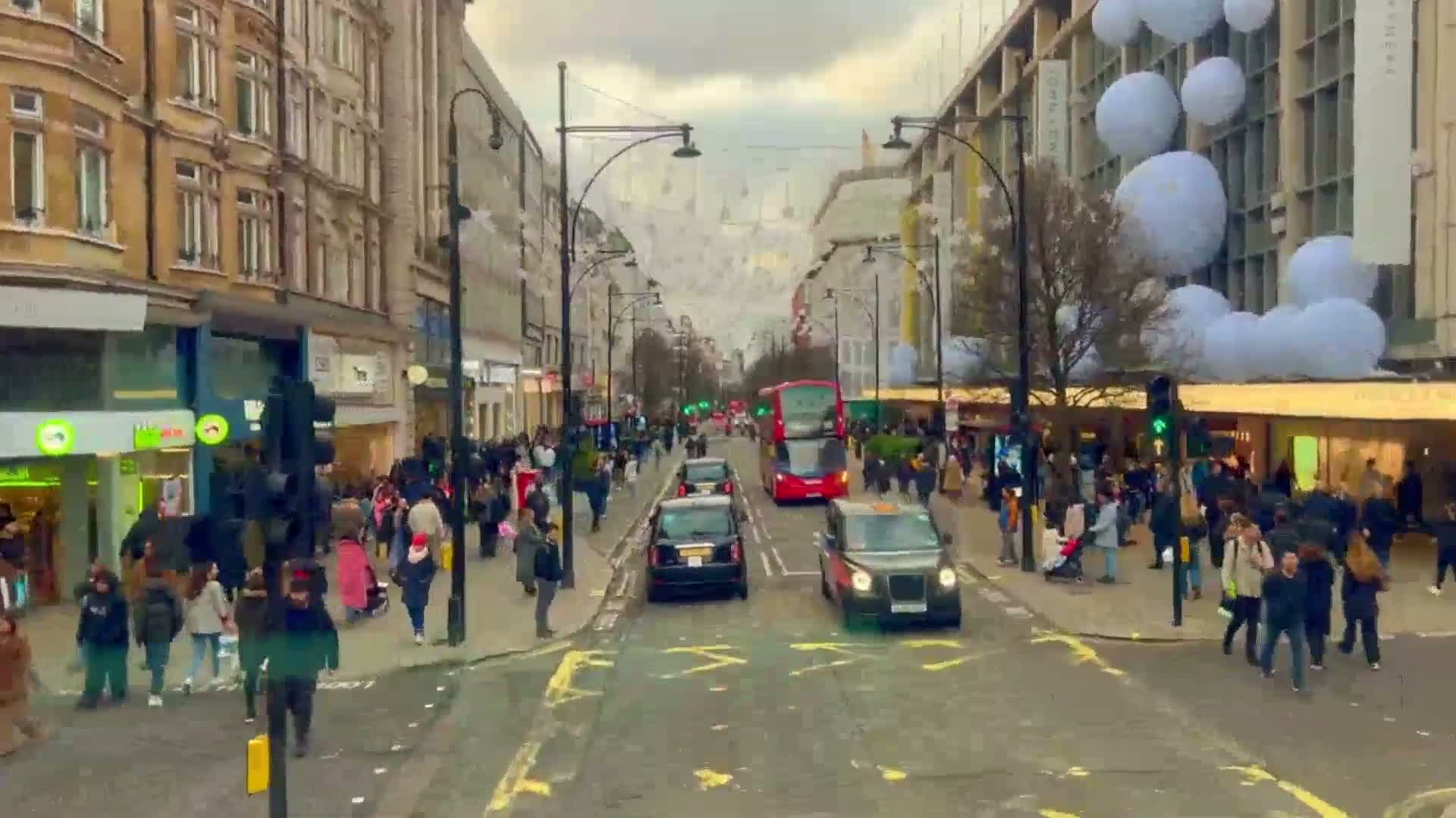 A busy city street with pedestrians and vehicles, including a double-decker bus, under an overcast sky, features large white balloons decorating buildings along the street.