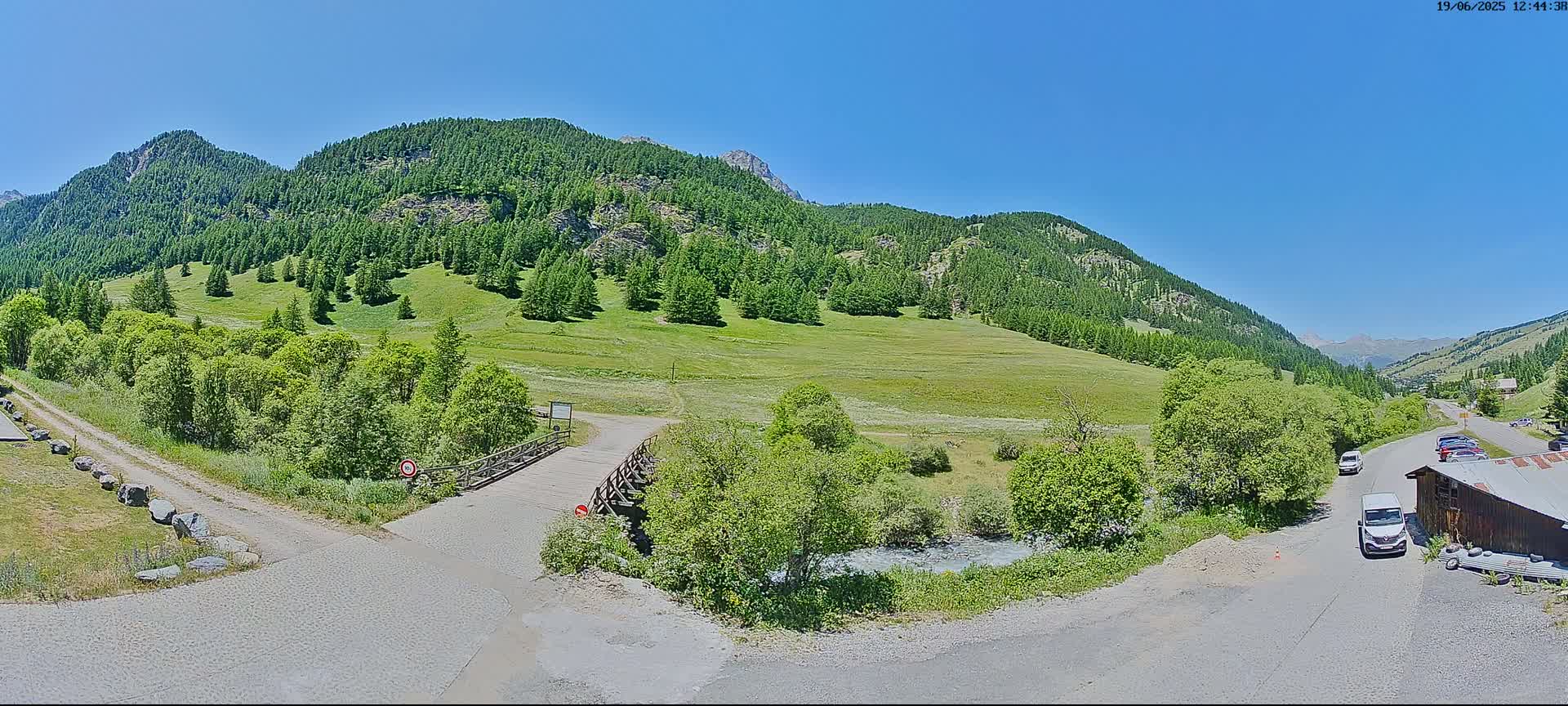 A mountain valley with green hillsides, lush vegetation, a small wooden bridge, and a gravel road under sunny skies.
