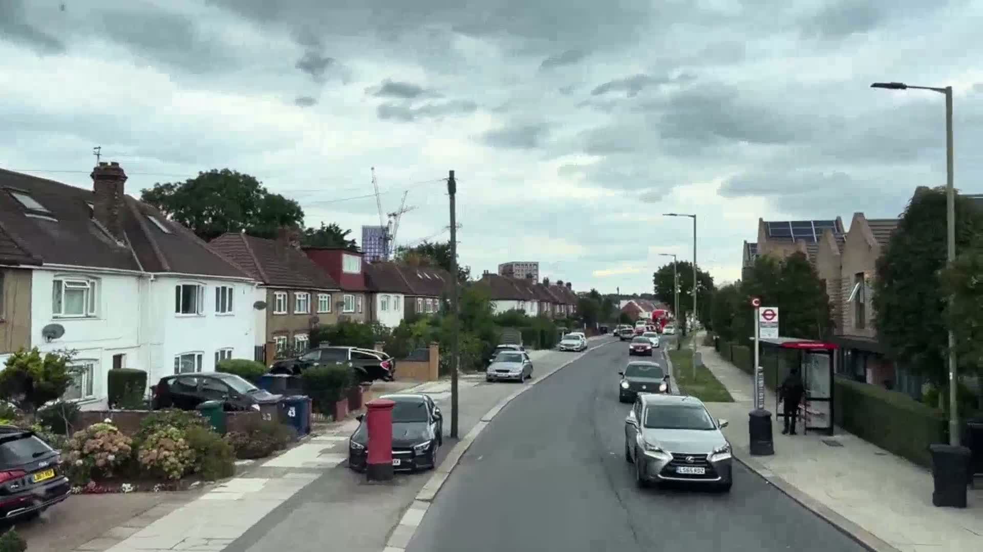 A residential street with parked cars and houses on either side under a cloudy sky, and a few cars driving down the road.
