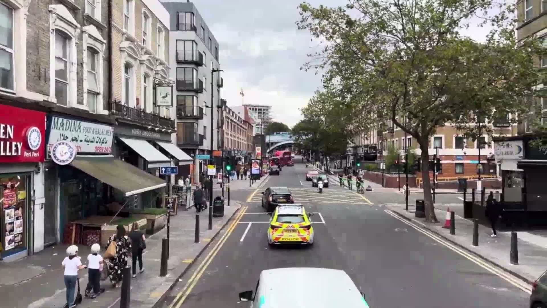 A police car drives down a city street lined with shops and buildings under an overcast sky.