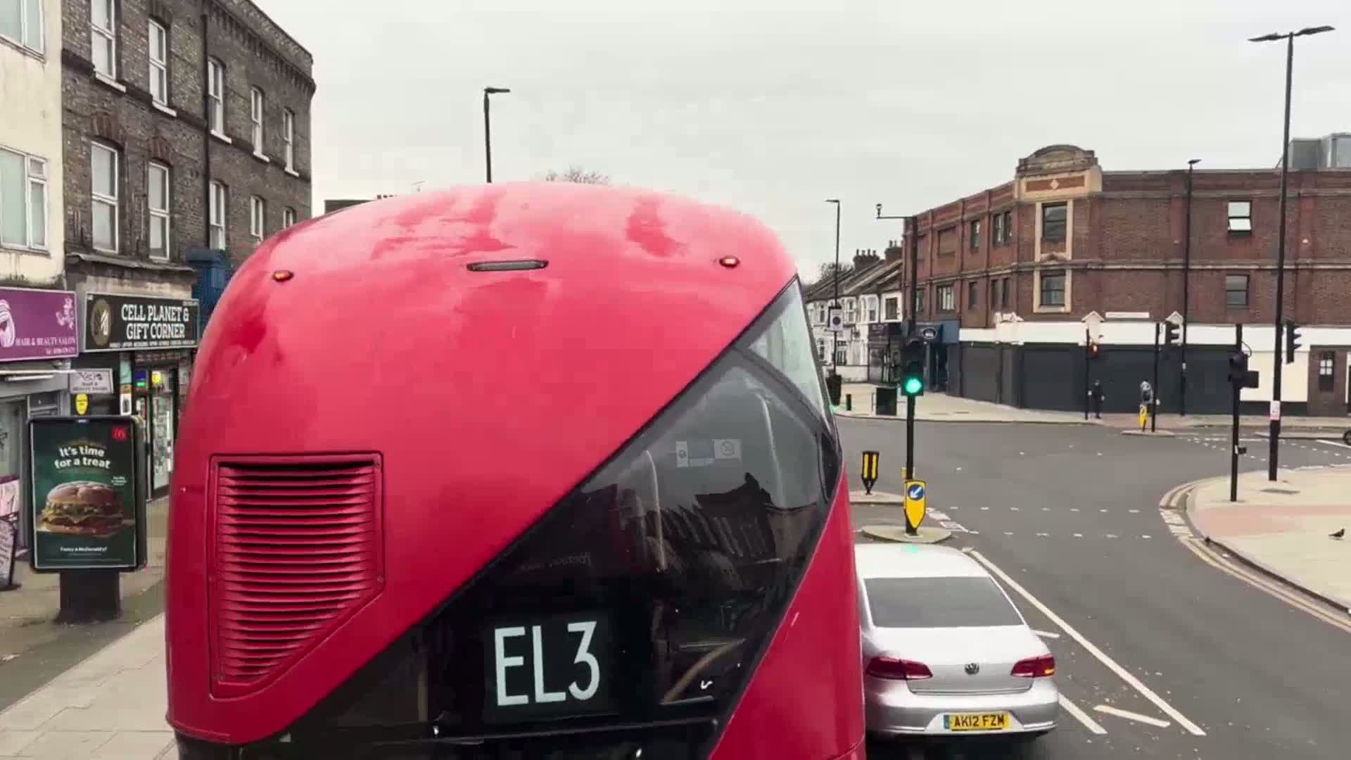 A red double-decker bus's front is visible, stopped at a traffic intersection on an overcast day, with a silver car visible behind it.