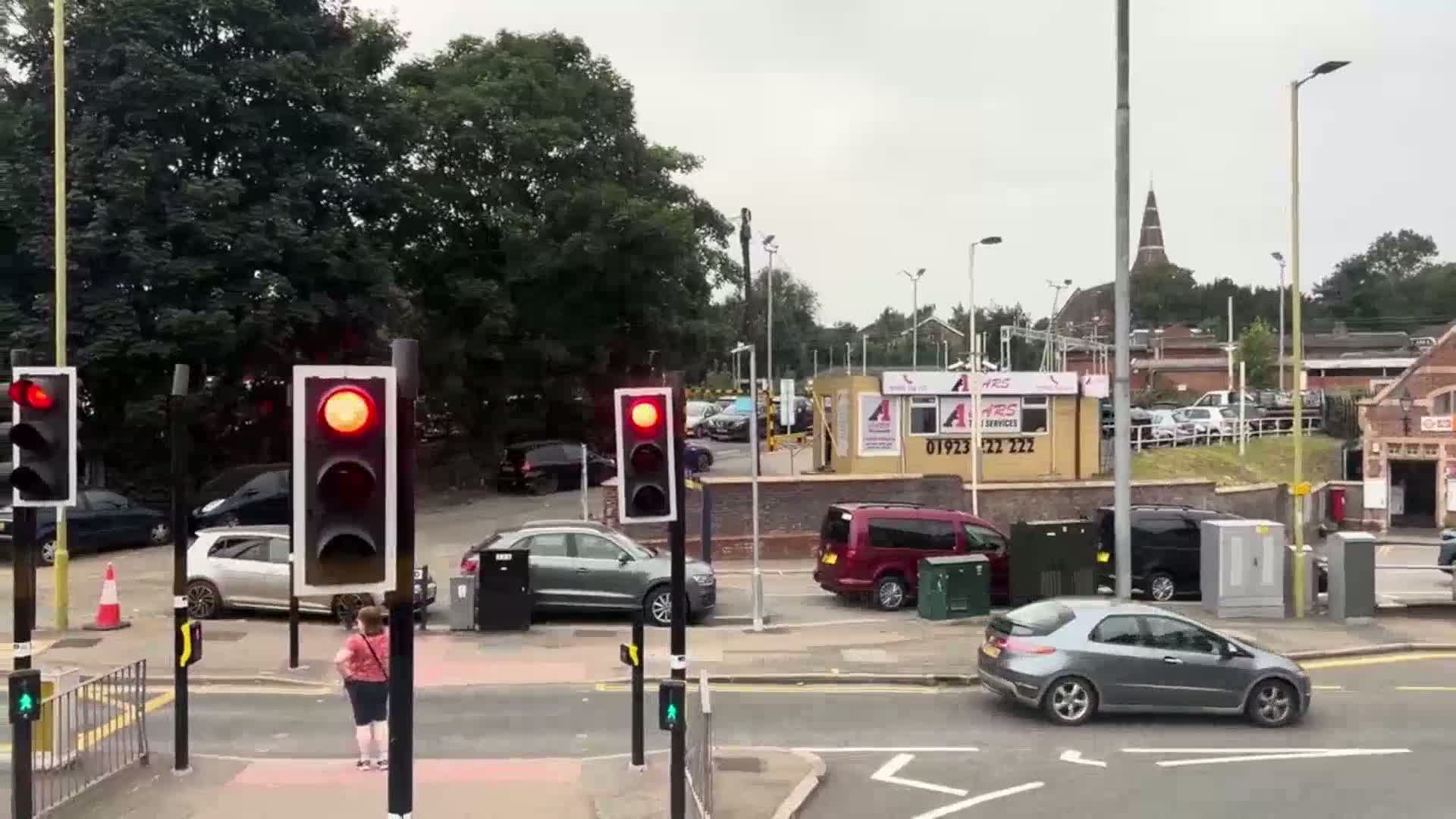 A street scene on an overcast day shows a pedestrian crossing at a red light, several cars stopped at a traffic light, and a parking lot with additional vehicles and a small building in the background.