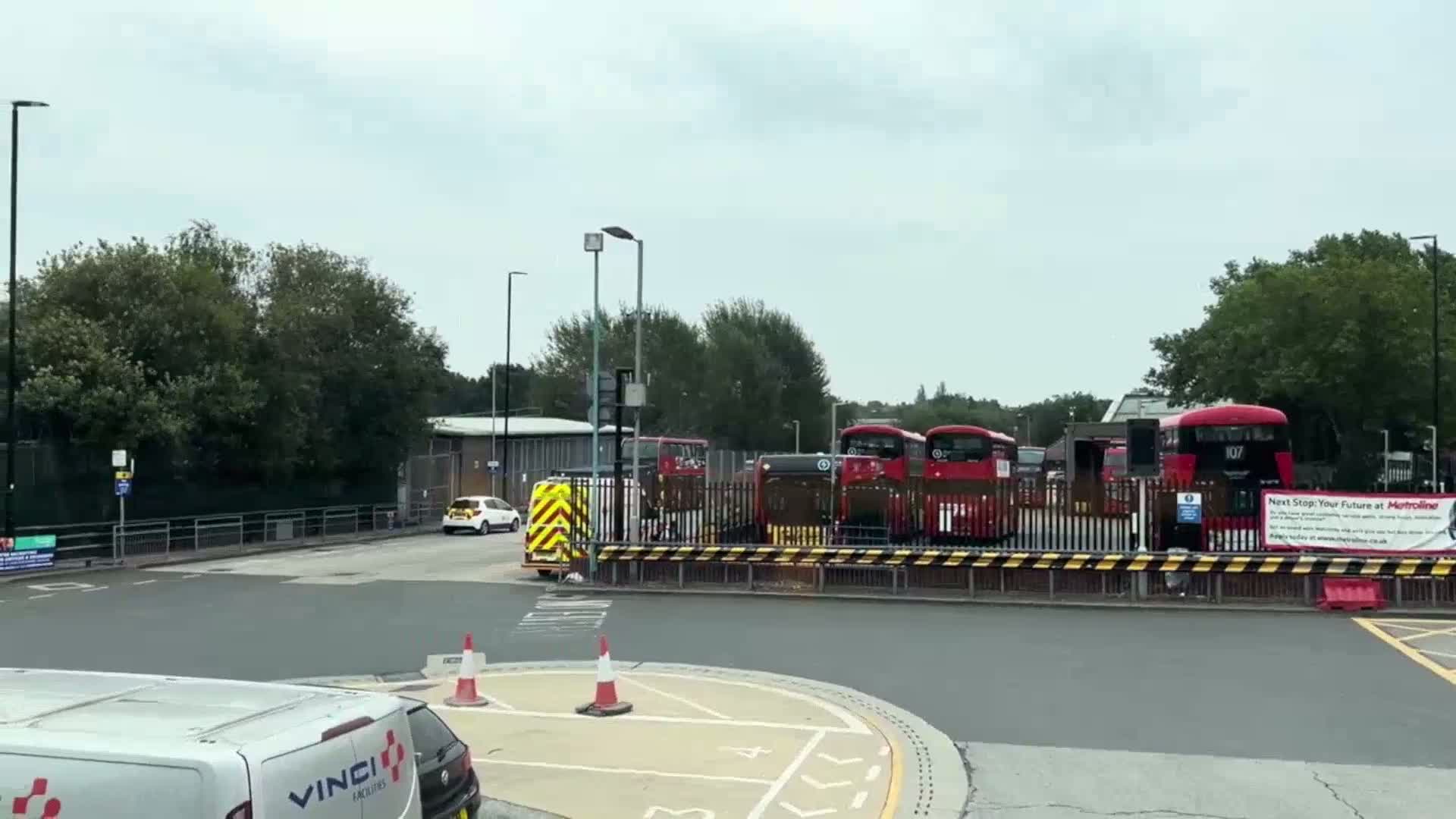 Several double-decker buses are parked in a depot on an overcast day, with a white van and a small car visible in the foreground.