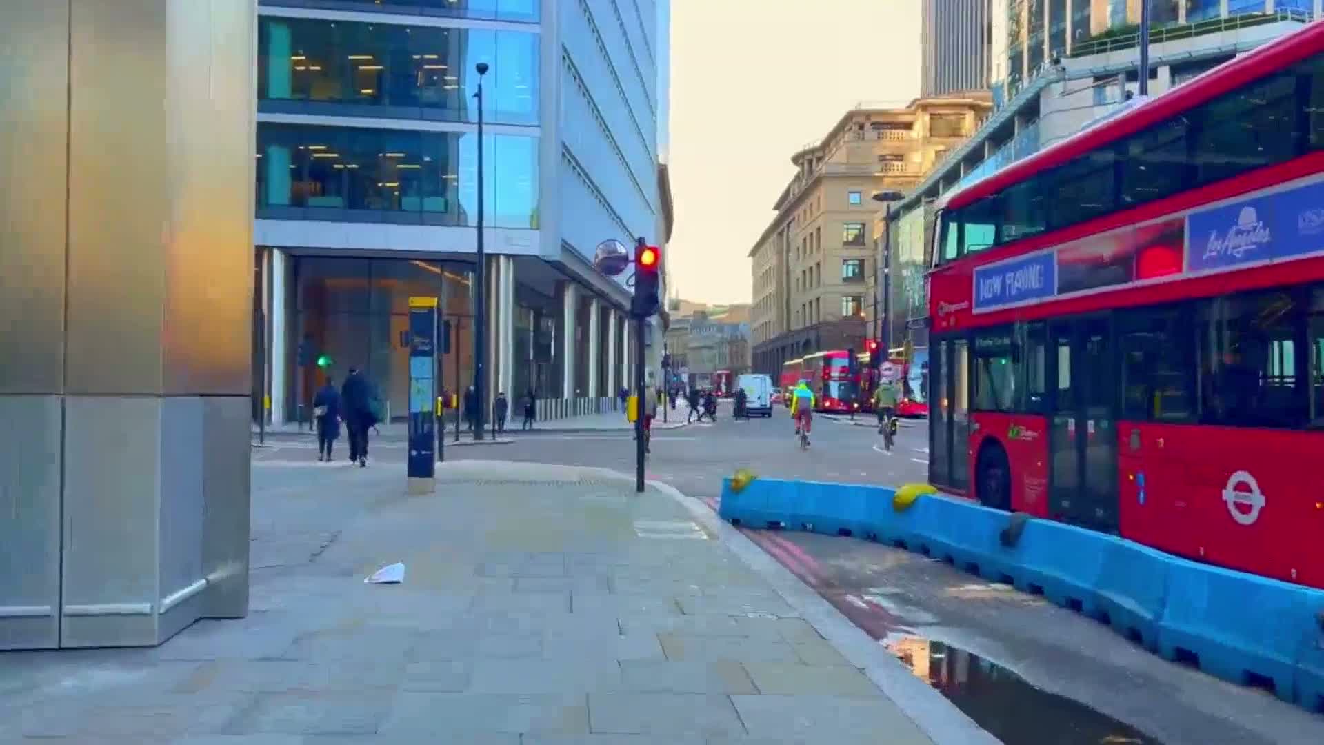 A city street scene on a bright, clear day shows a red double-decker bus stopped at a traffic light, pedestrians walking on the sidewalk, and cyclists riding in the street.