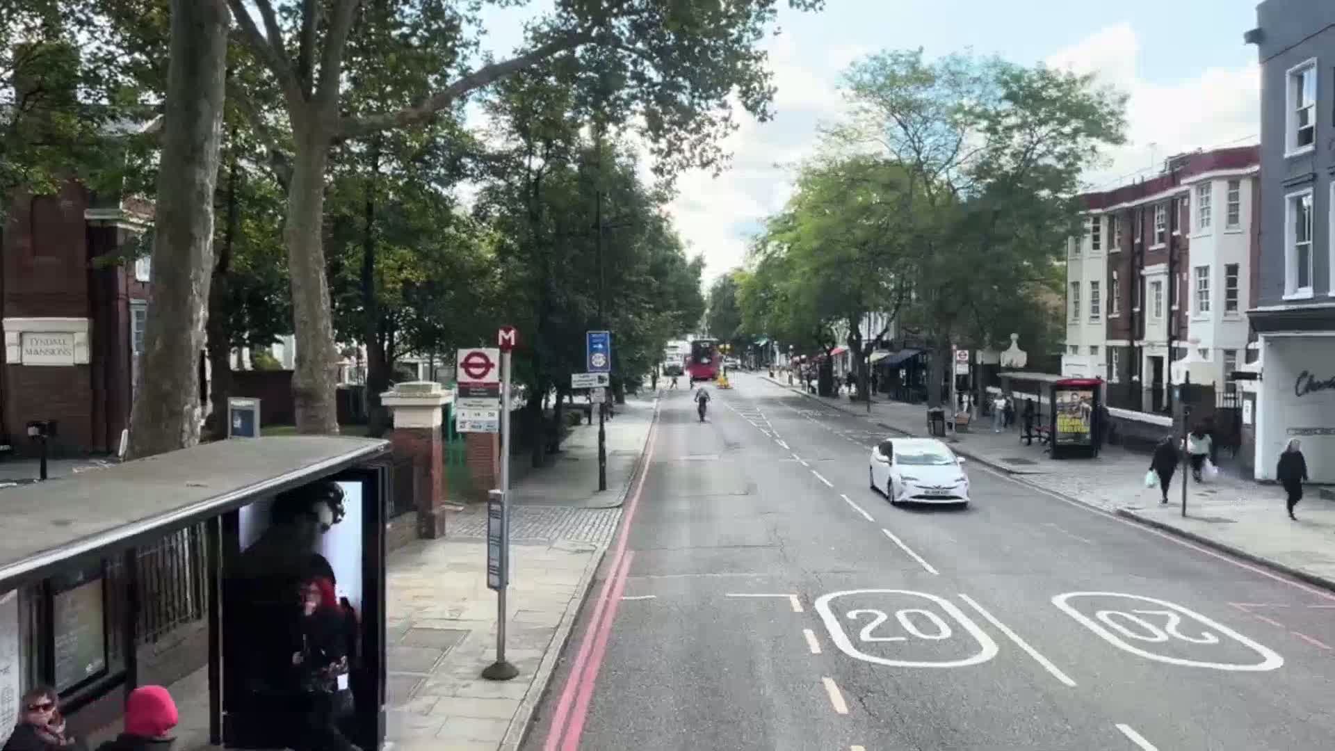 A city street scene on a bright, clear day shows a red double-decker bus stopped at a traffic light, pedestrians walking on the sidewalk, and cyclists riding in the street.