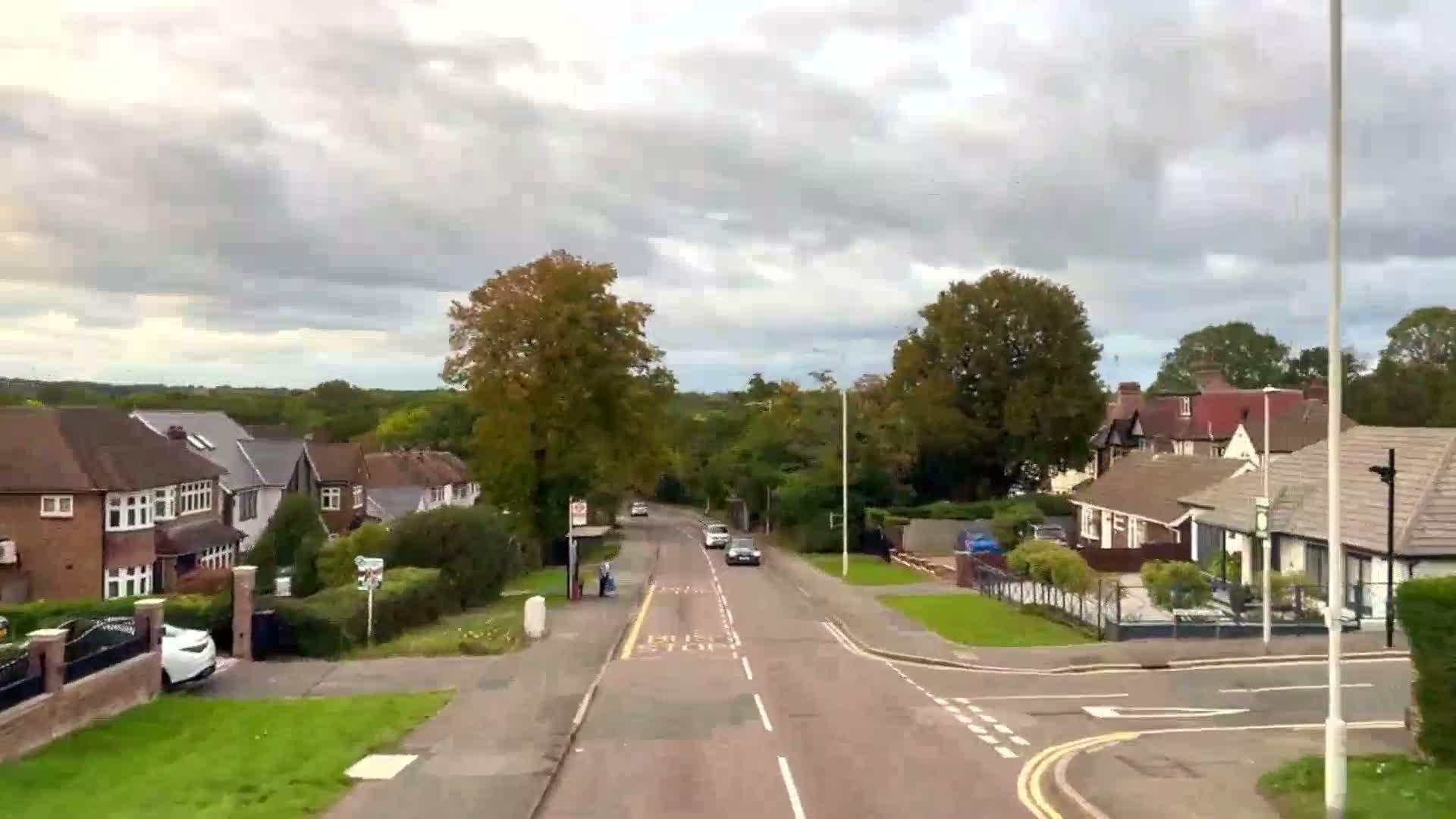 A suburban street lined with houses and numerous trees, featuring a road with several cars and a bus stop, stretches under a heavily overcast sky.
