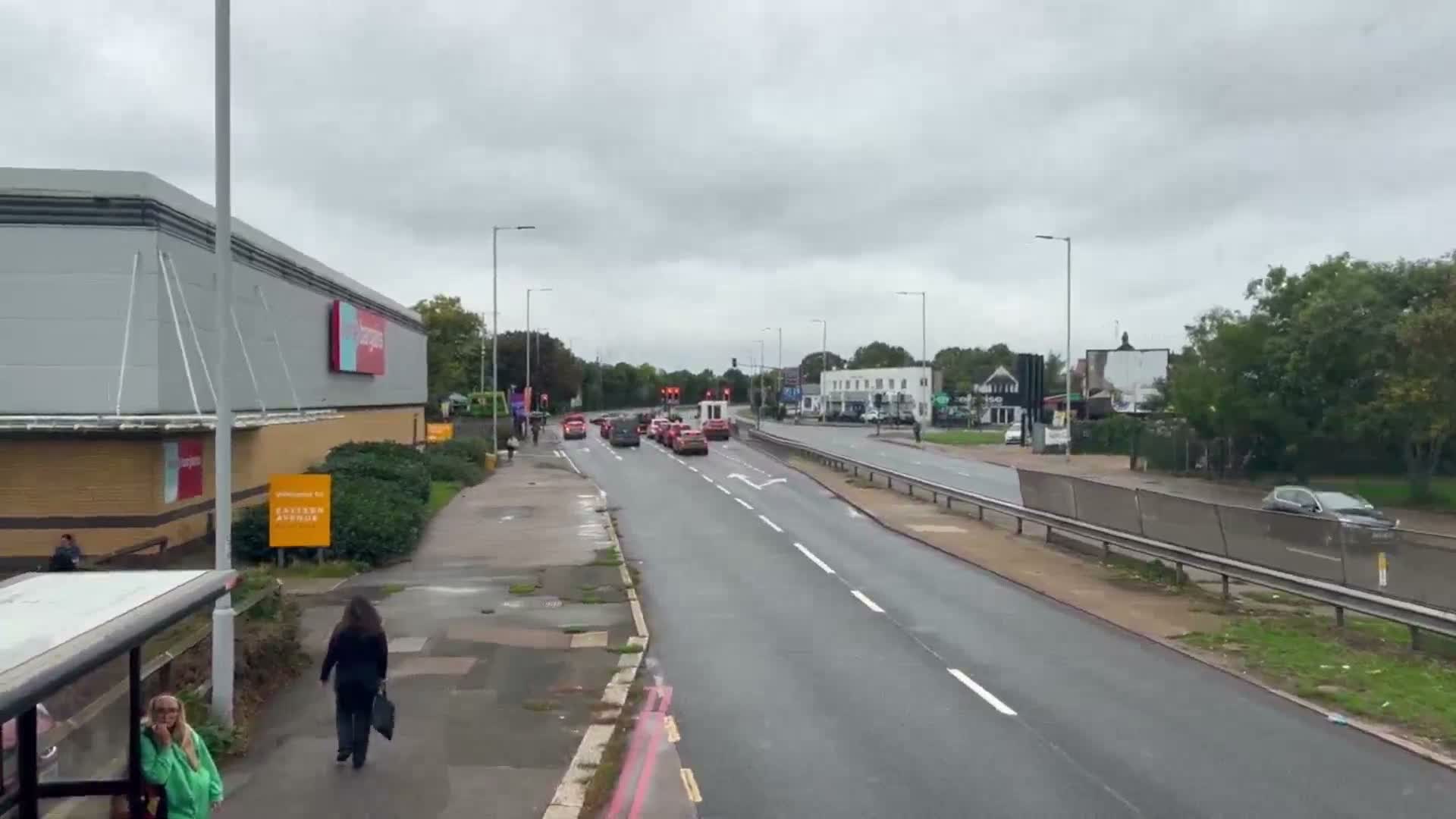 A suburban street lined with houses and numerous trees, featuring a road with several cars and a bus stop, stretches under a heavily overcast sky.