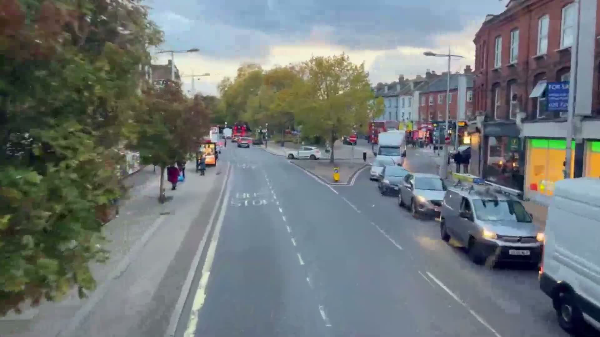 An elevated view captures a bustling urban street on an overcast day, featuring a thoroughfare with moving vehicles and parked cars, sidewalks populated by pedestrians, and a backdrop of brick buildings and green trees.