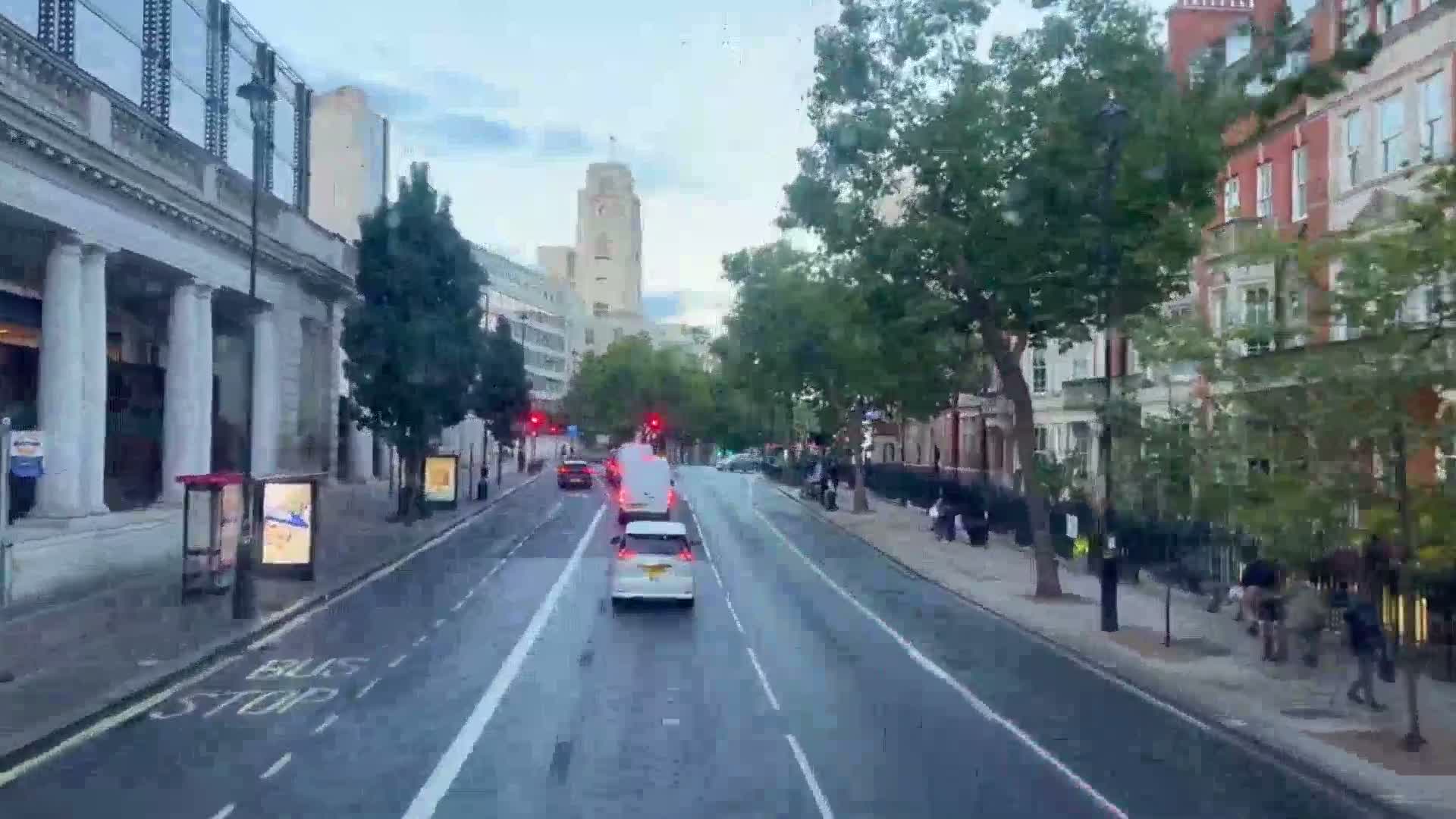 An elevated view captures a bustling urban street on an overcast day, featuring a thoroughfare with moving vehicles and parked cars, sidewalks populated by pedestrians, and a backdrop of brick buildings and green trees.