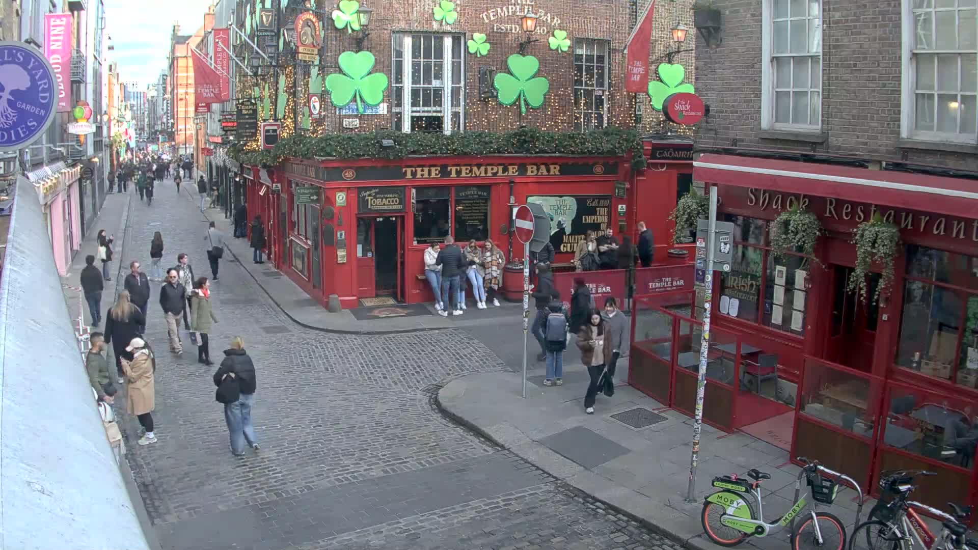 A cobblestone street scene shows several people walking past red brick buildings, one of which is a pub with shamrock decorations, under an overcast sky.