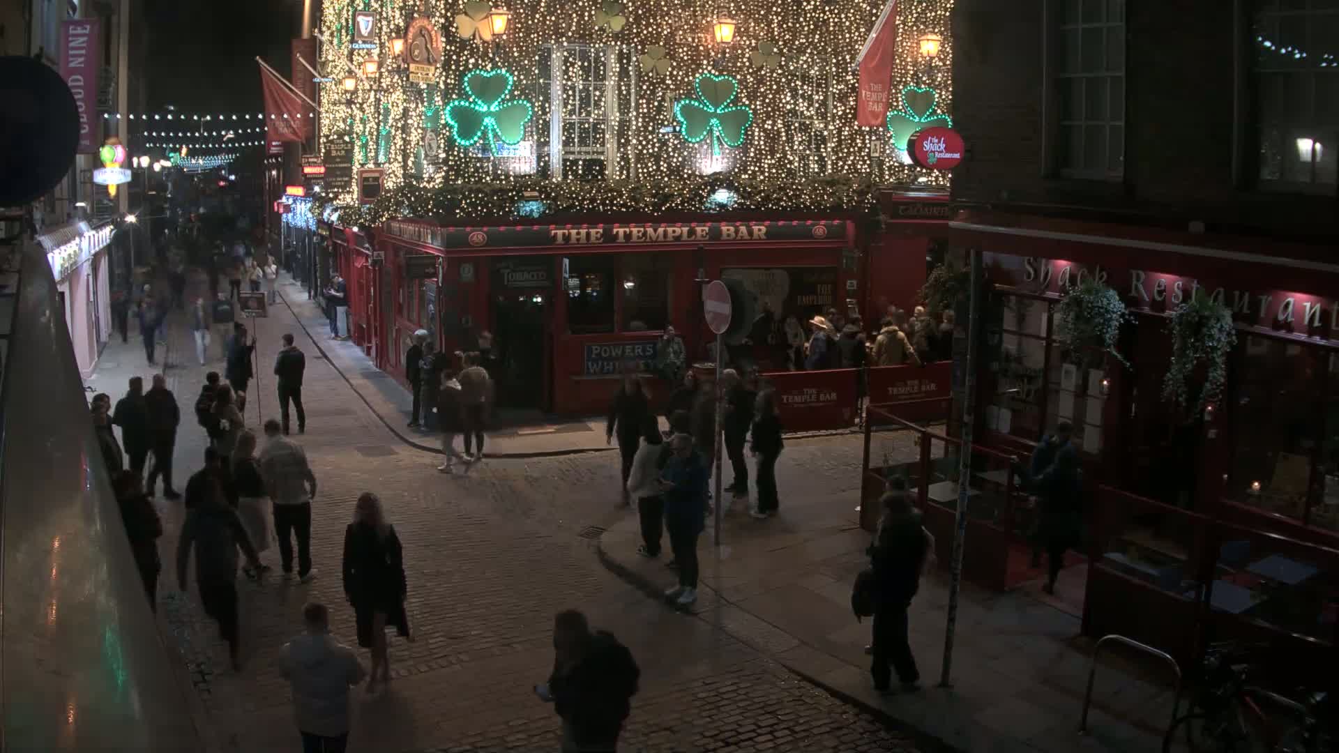 A nighttime street scene shows many people walking around a cobblestone street in front of a brightly lit pub decorated with shamrocks and strings of lights.