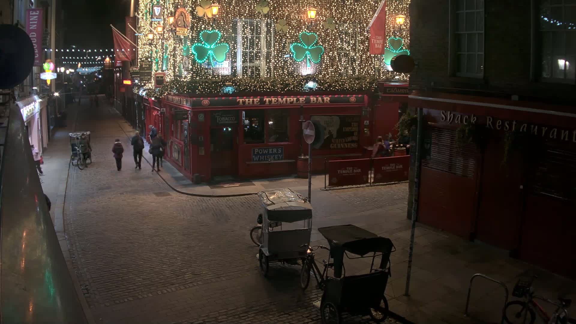 A cobblestone street at night, illuminated by streetlights and the bright lights of a decorated pub, shows several people walking and two rickshaws parked.