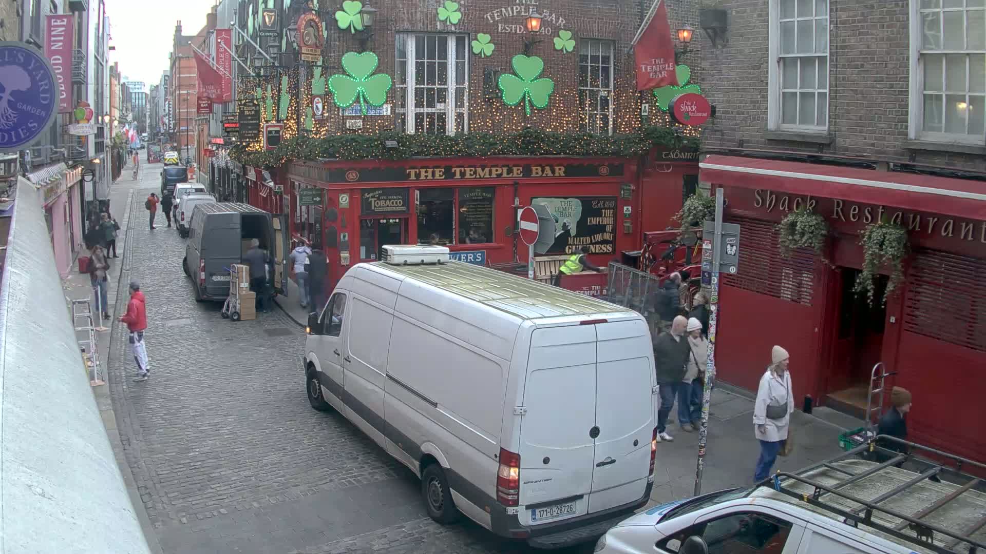 A cobblestone street, under an overcast sky, shows several people walking past delivery vans and a red pub decorated with shamrocks and lights.
