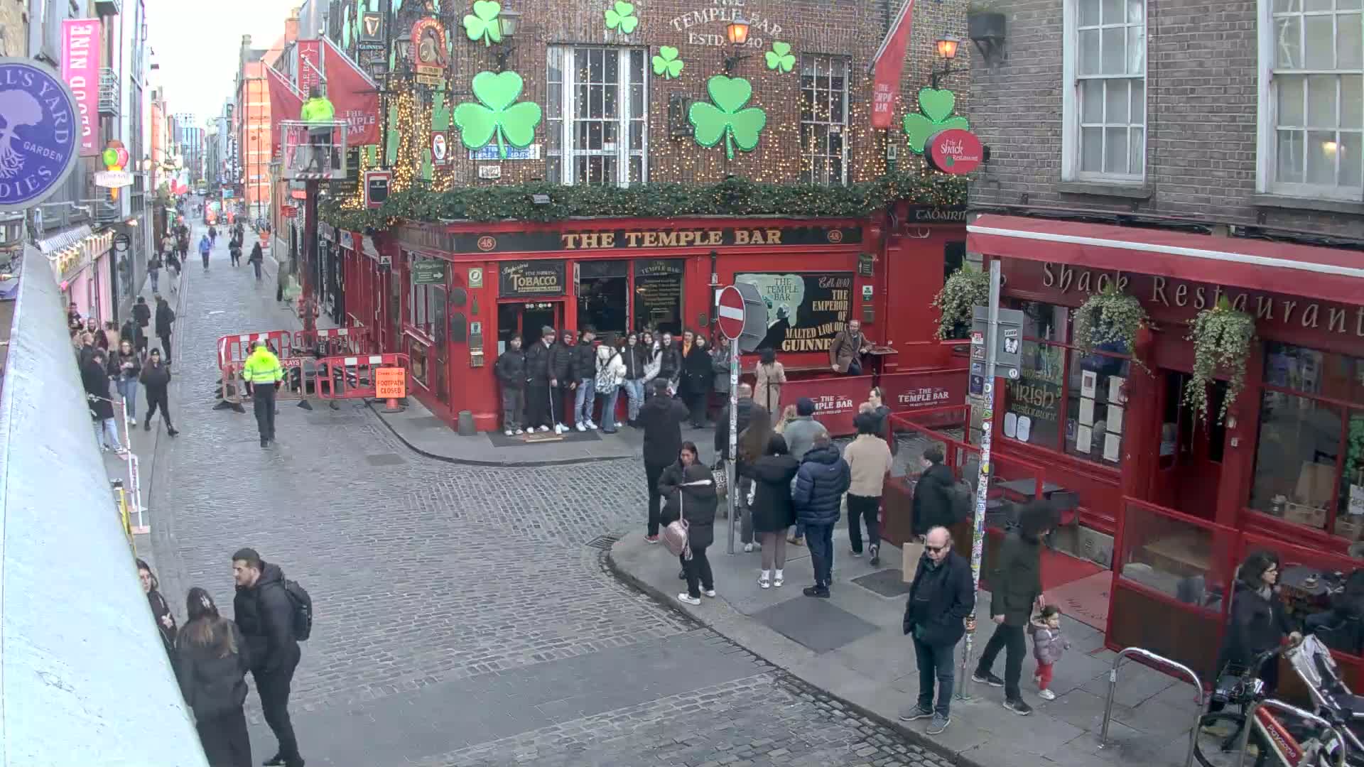 A cobblestone street, possibly in a European city, is populated with pedestrians and features red brick buildings adorned with shamrocks and string lights, under an overcast sky.
