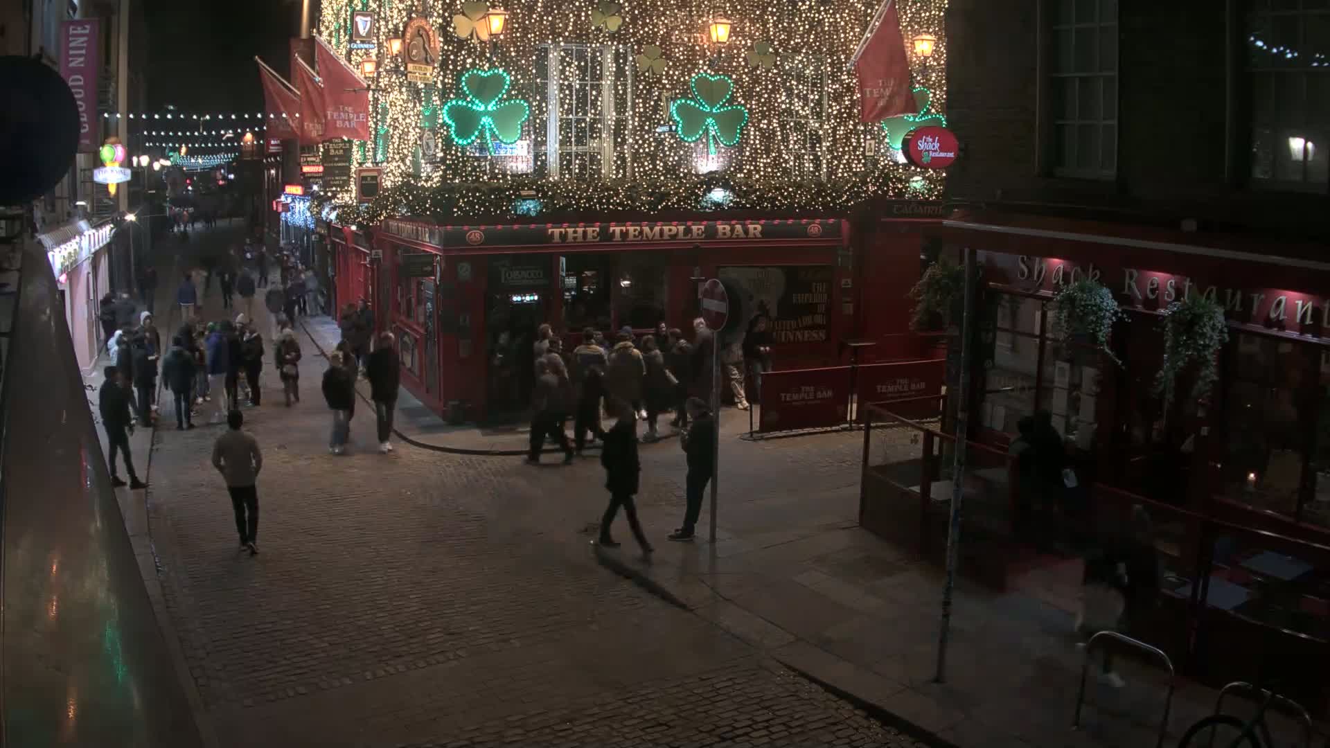A nighttime street scene shows a cobblestone street with many pedestrians walking past a brightly lit pub decorated with shamrocks and lights.
