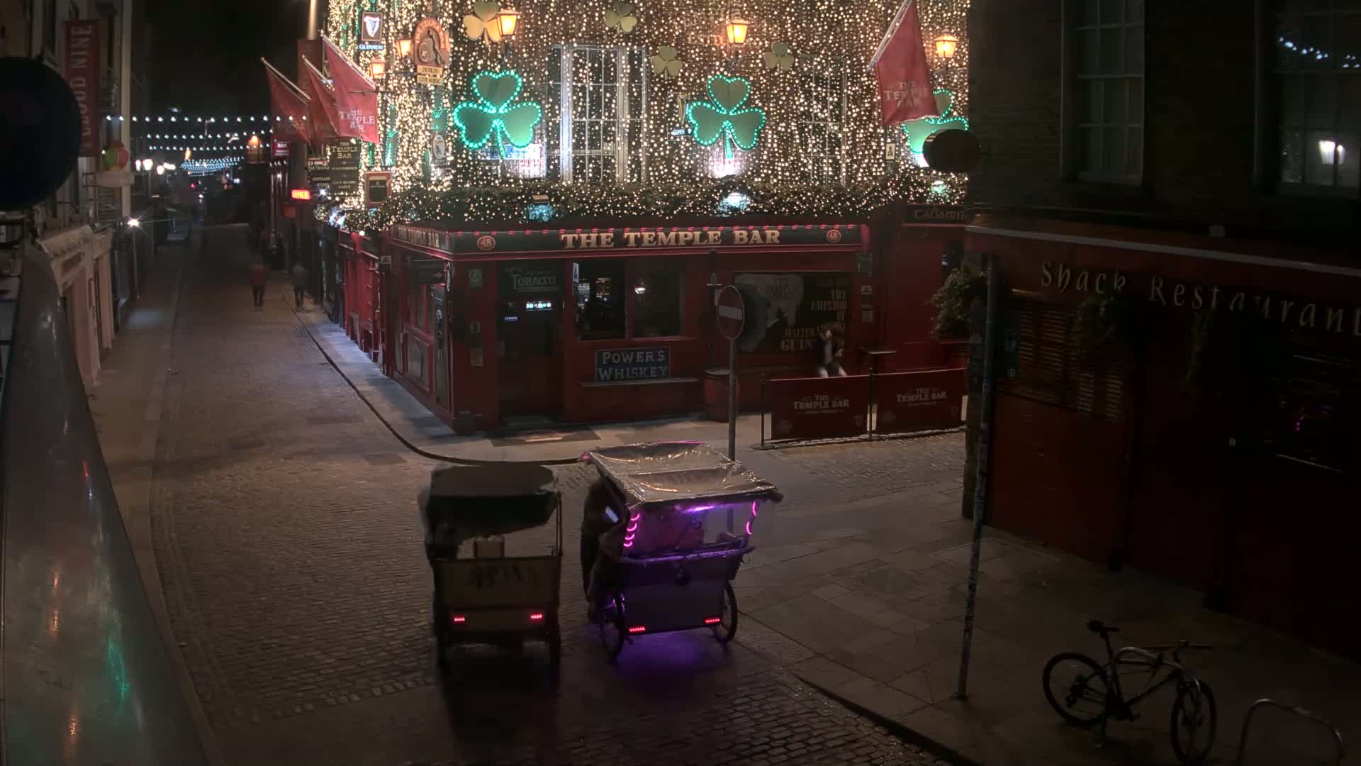 At night, two rickshaws are parked on a cobblestone street in front of a brightly lit pub decorated with shamrocks and string lights.