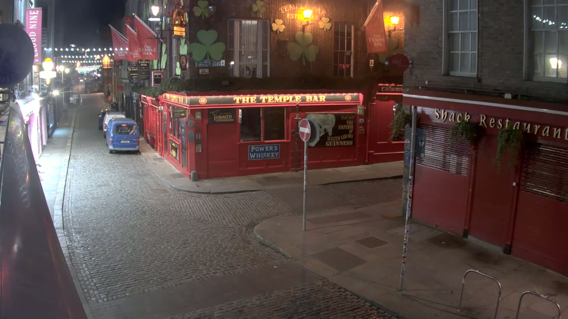 A cobblestone street at night, illuminated by streetlights, shows a red-brick building with a pub and restaurant, and a few parked vehicles.